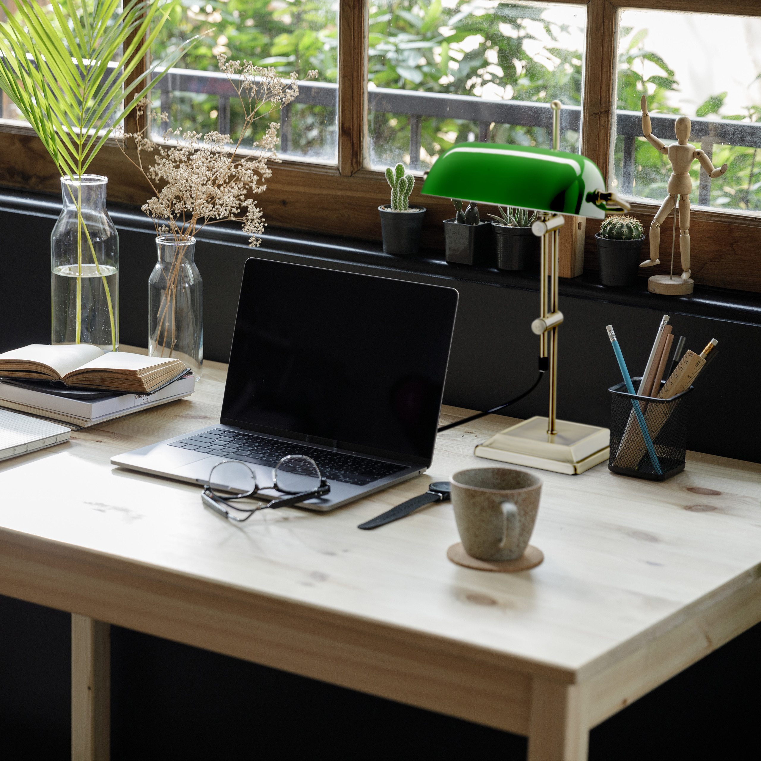 Ein Holzschreibtisch mit Laptop, grüner Lampe, Büchern, Brille, Tasse und Pflanzen vor einem Fenster.