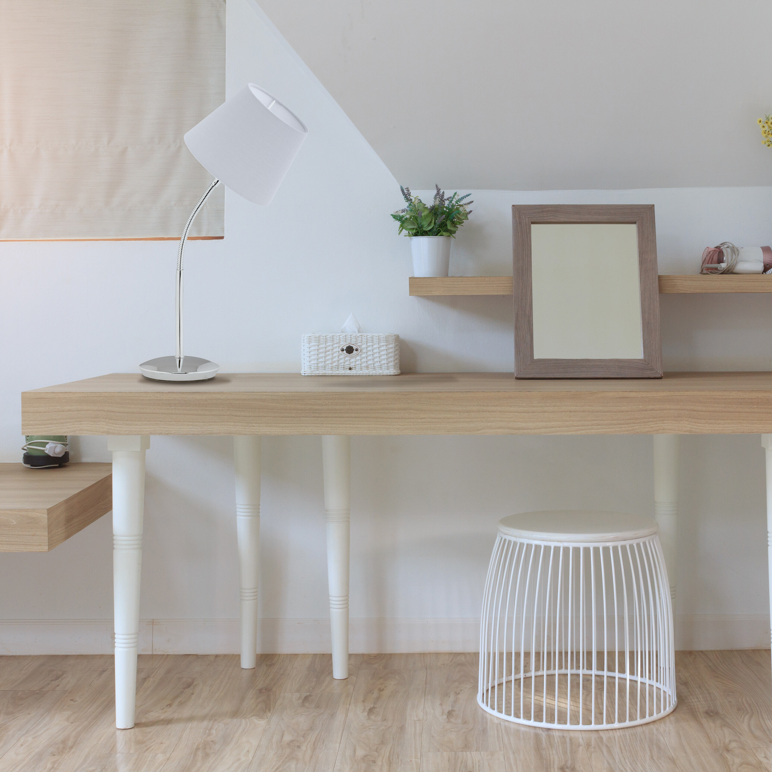 Ein Holztisch mit weißer Lampe, Box, Rahmen und weißem DrahtHocker vor einer weißen Wand.