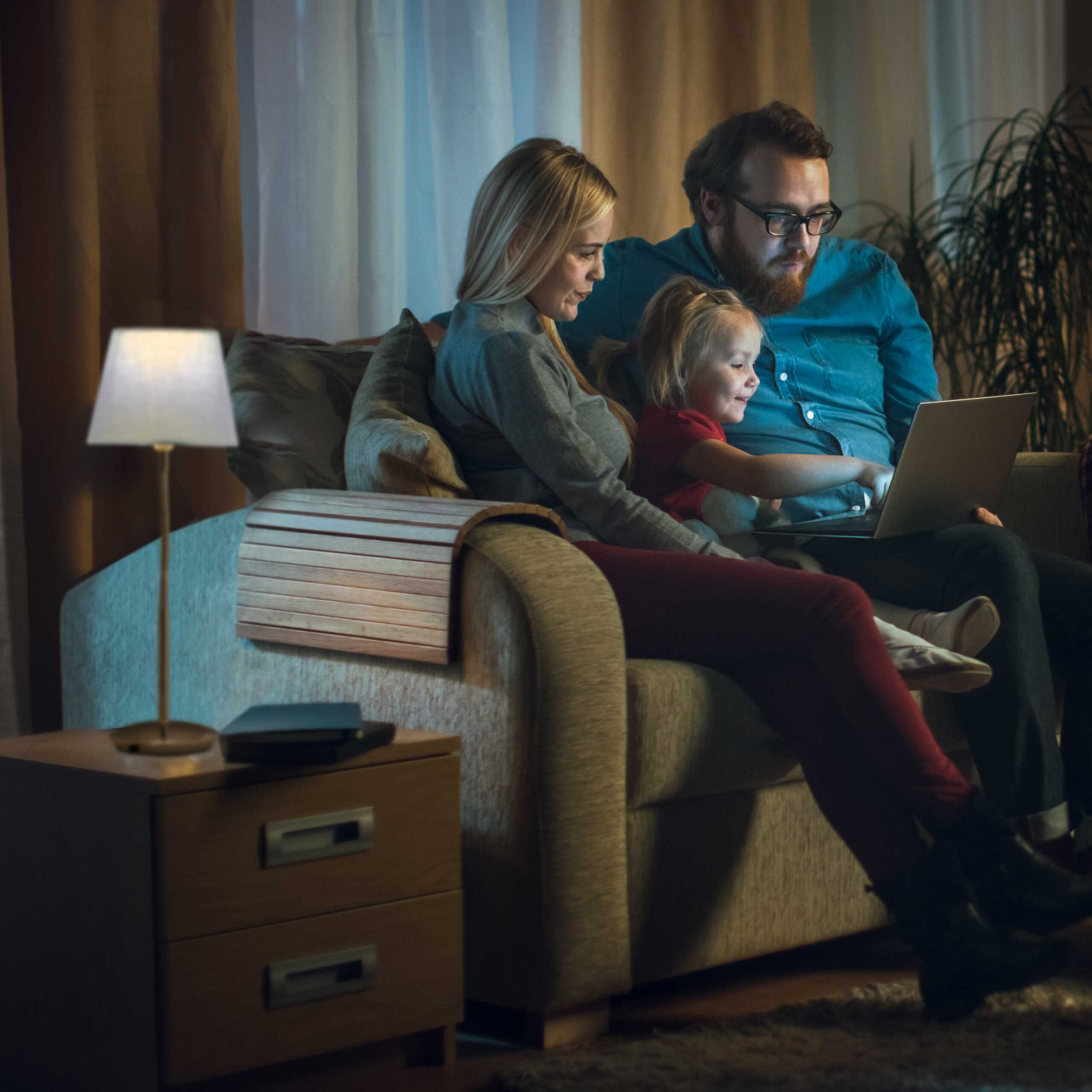 Eine dreiköpfige Familie sitzt in einem gedimmten Raum auf einem Sofa und schaut auf einen Laptop.