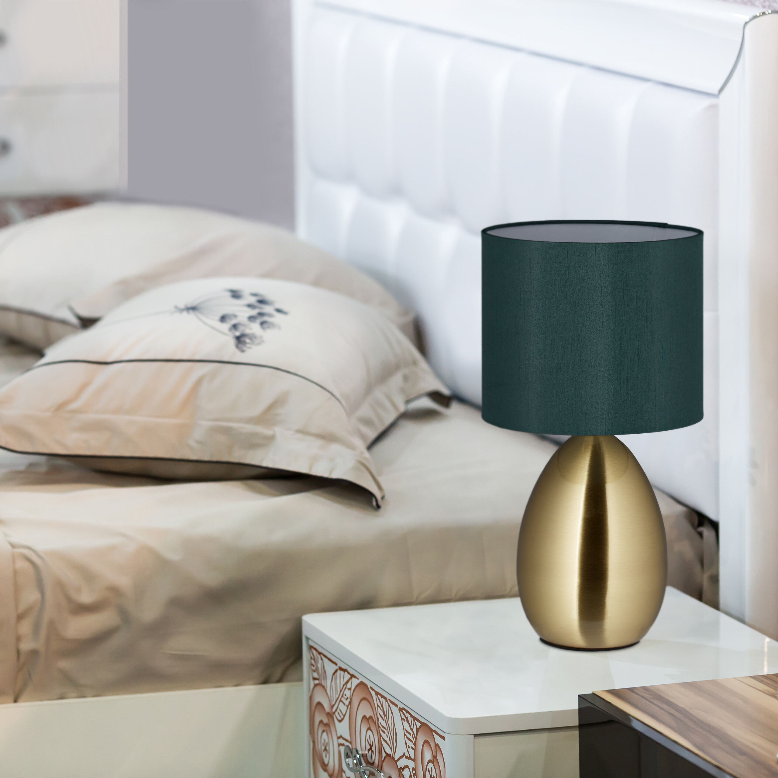 Eine grüne Tischlampe mit goldenem Fuß steht auf einem Nachttisch neben einem Bett mit gemusterten Kissen.