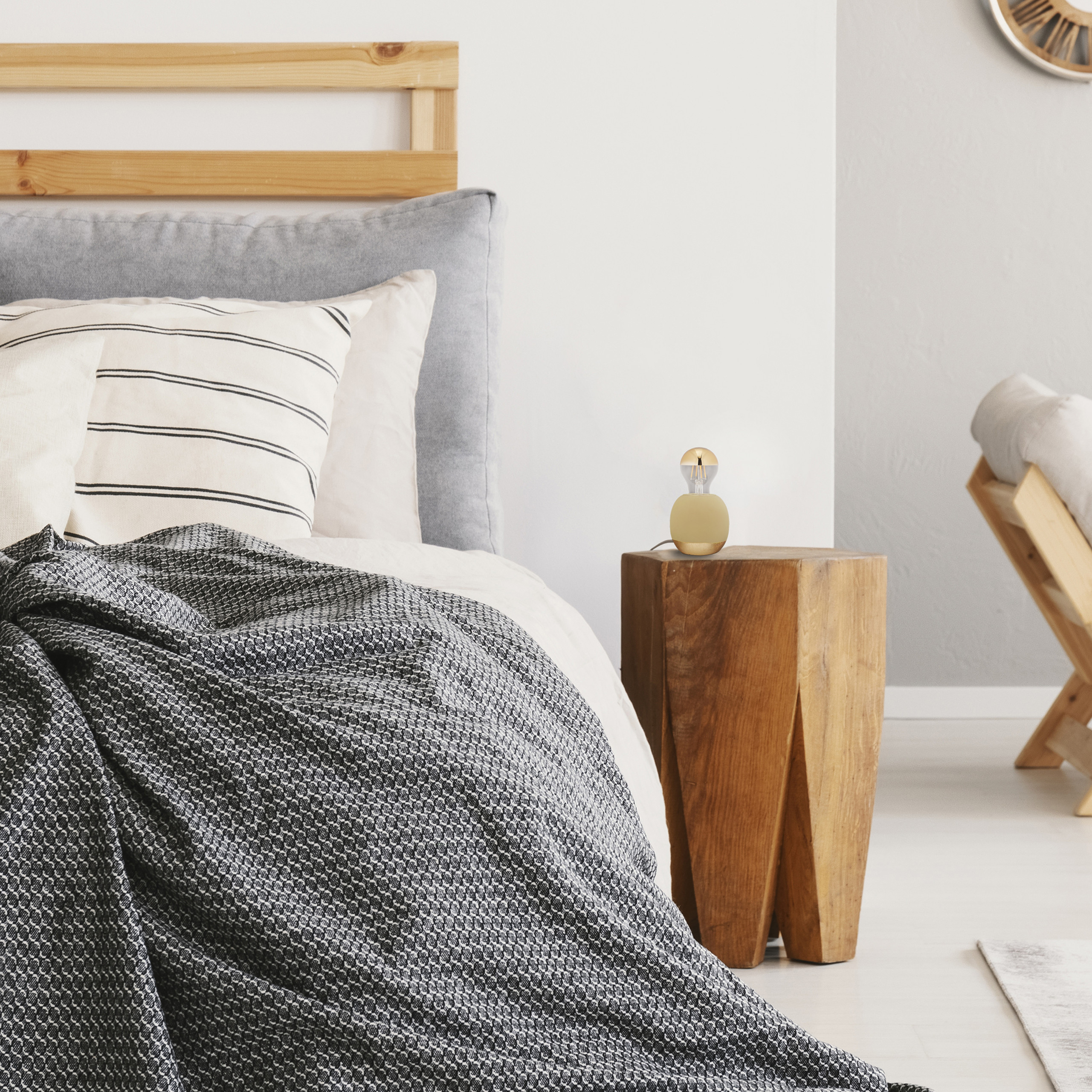 Ein Bett mit grauem Kopfteil, gemusterten Kissen und einer grauen Decke, daneben ein Holztisch mit Lampe.