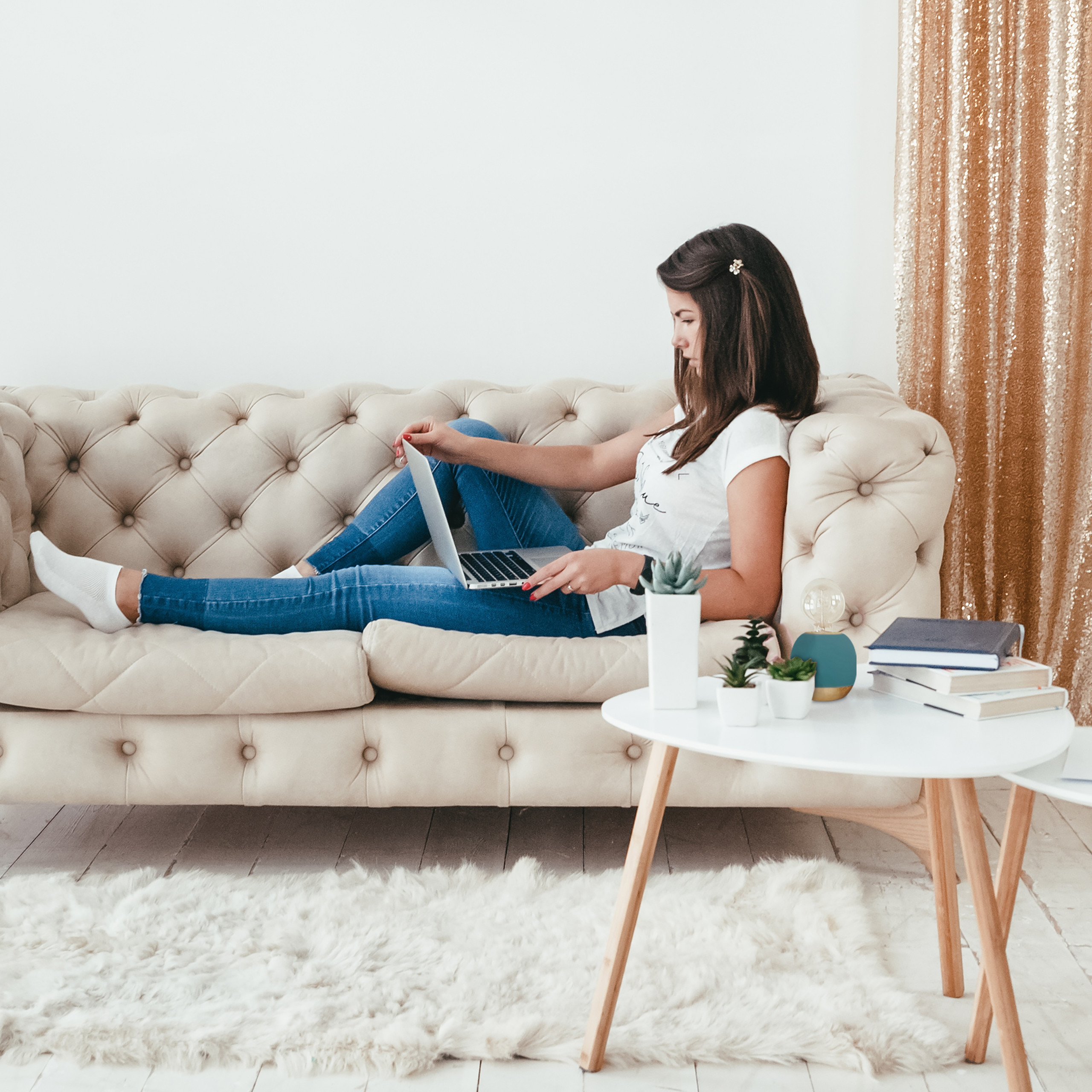 Eine Frau in Jeans und weißem Shirt sitzt mit einem Laptop auf einem beigen Sofa neben einem Tisch.
