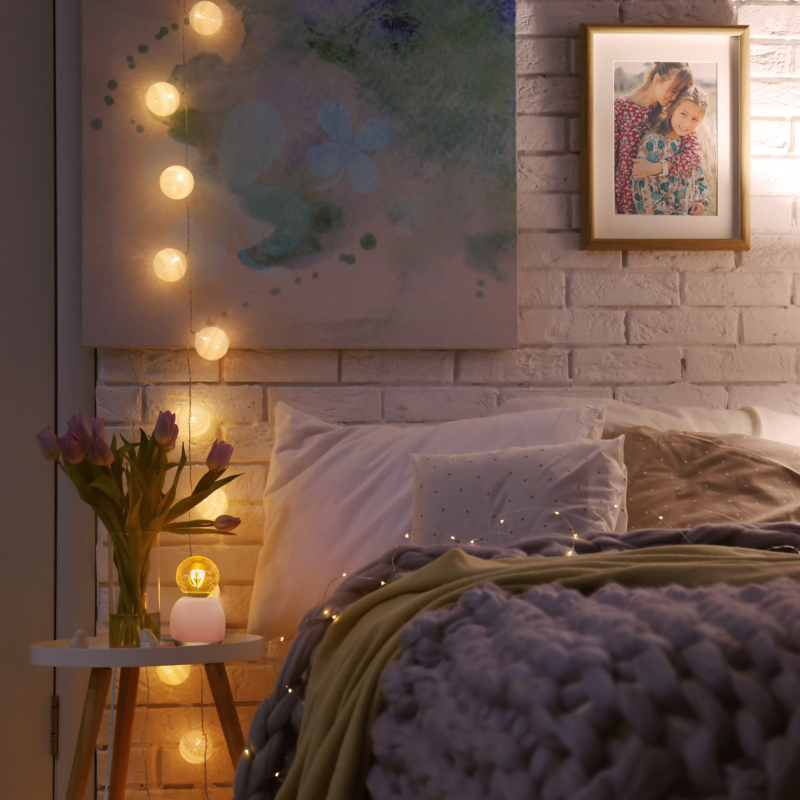Gemütliches Schlafzimmer mit weißer Backsteinwand, Lichterkette, Blumen in Vase und einem gerahmten Foto.