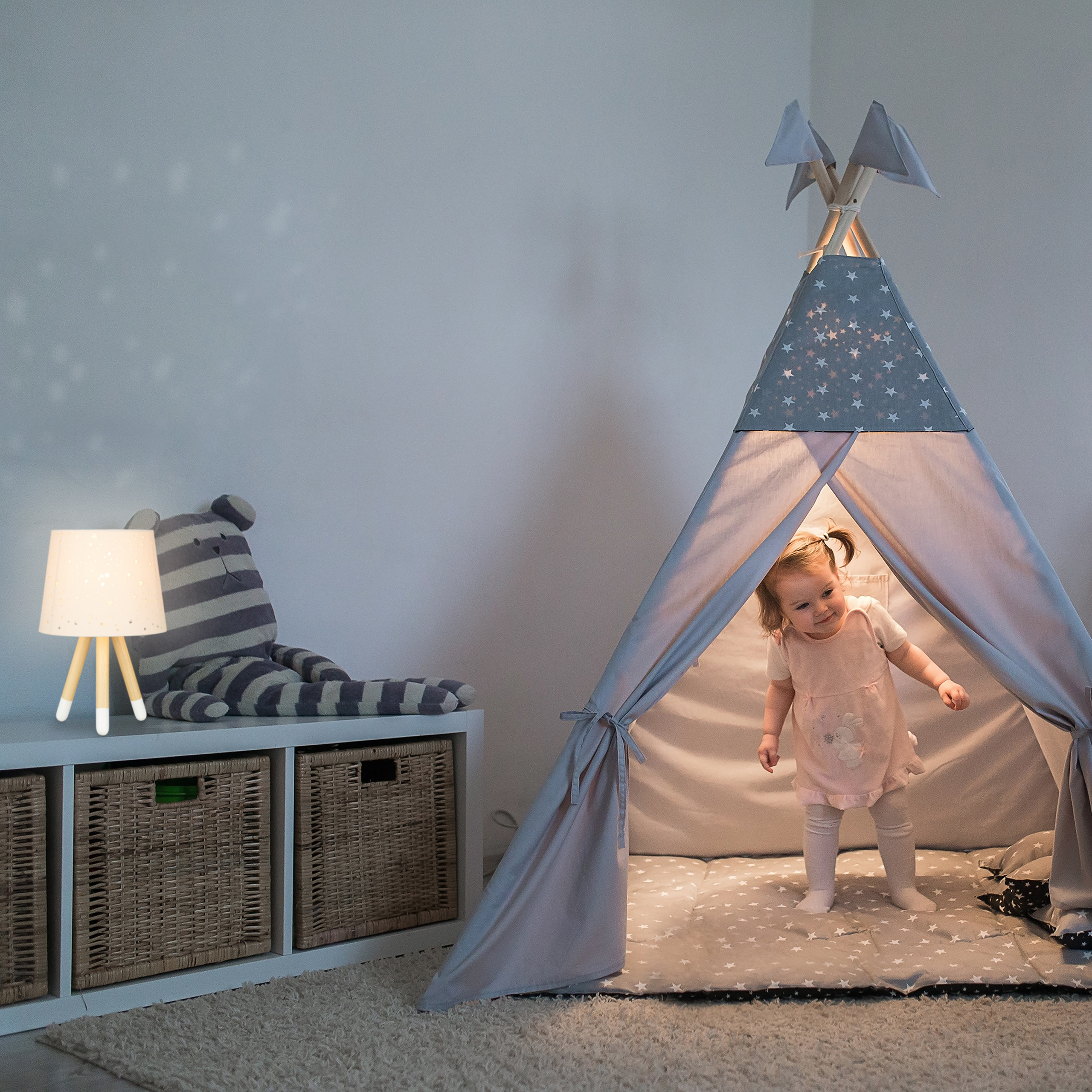 Kleines Mädchen in rosafarbenem Kleid steht in einem grauen Tipi-Zelt mit Sternenmuster, neben einem Stoffsitz.