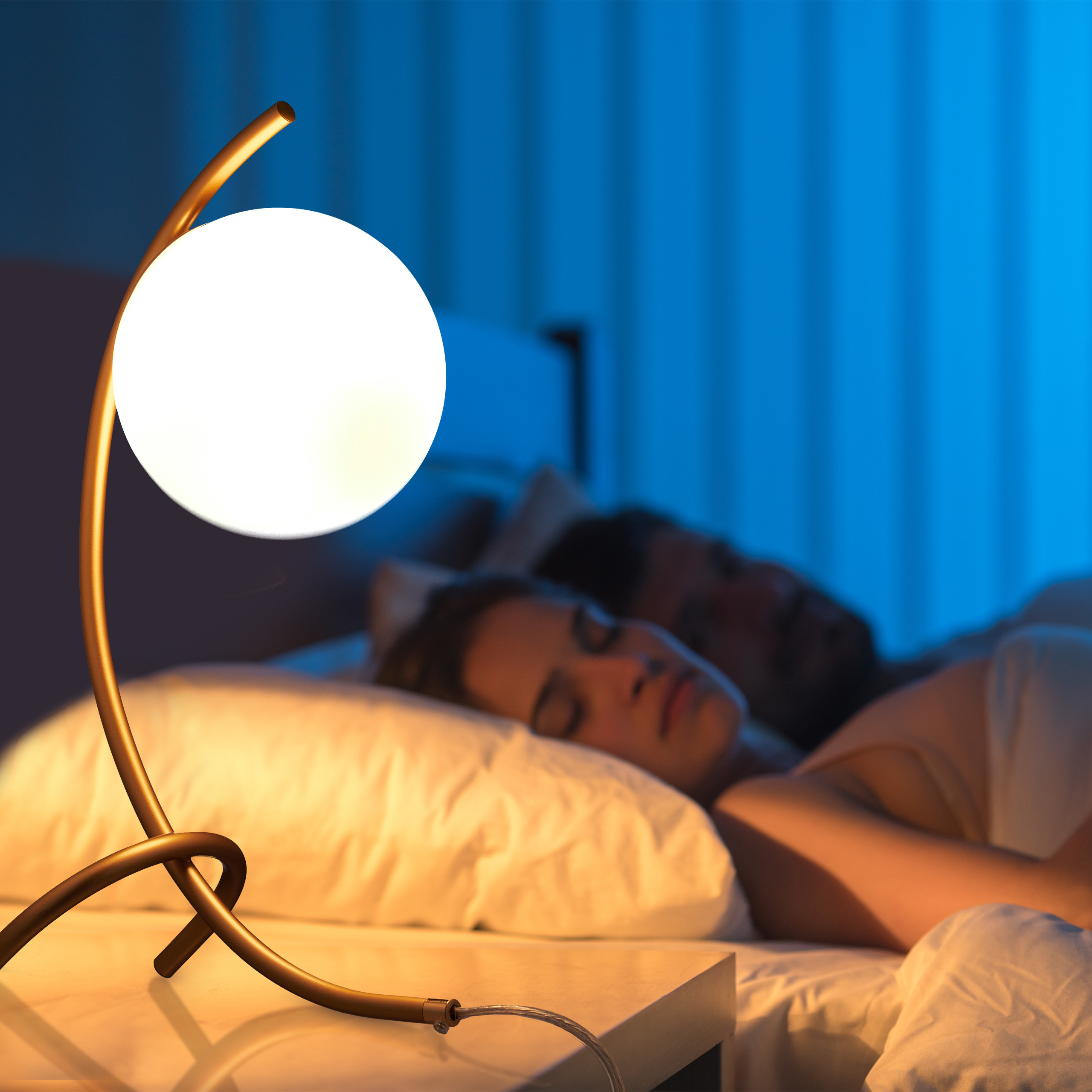 Eine moderne Nachttischlampe mit runder Glühbirne leuchtet, während ein Paar im Hintergrund schläft.