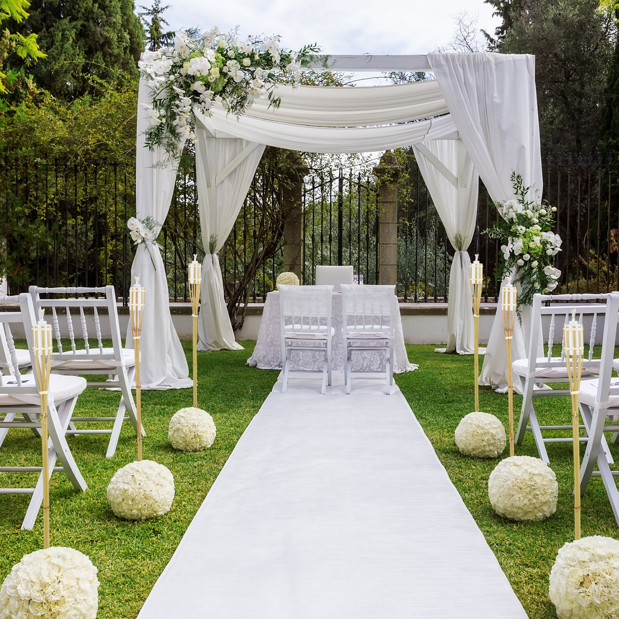 Eine weiße Hochzeitspergola im Freien mit weißem Teppich, Stühlen, Blumen und Fackeln auf der Rasenfläche.