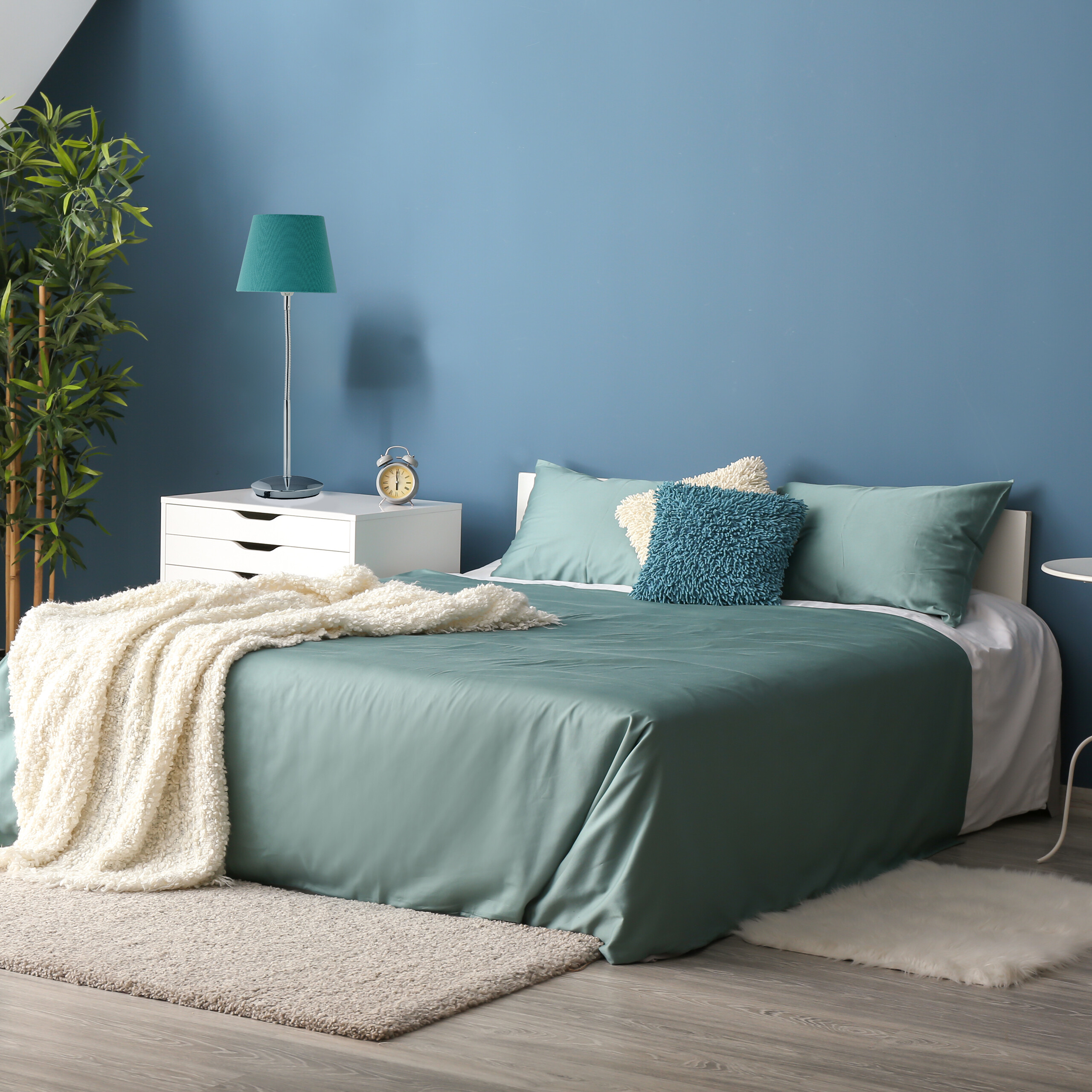 Ein Schlafzimmer mit blauem Bett, Nachttisch, Lampe und Zimmerpflanze.