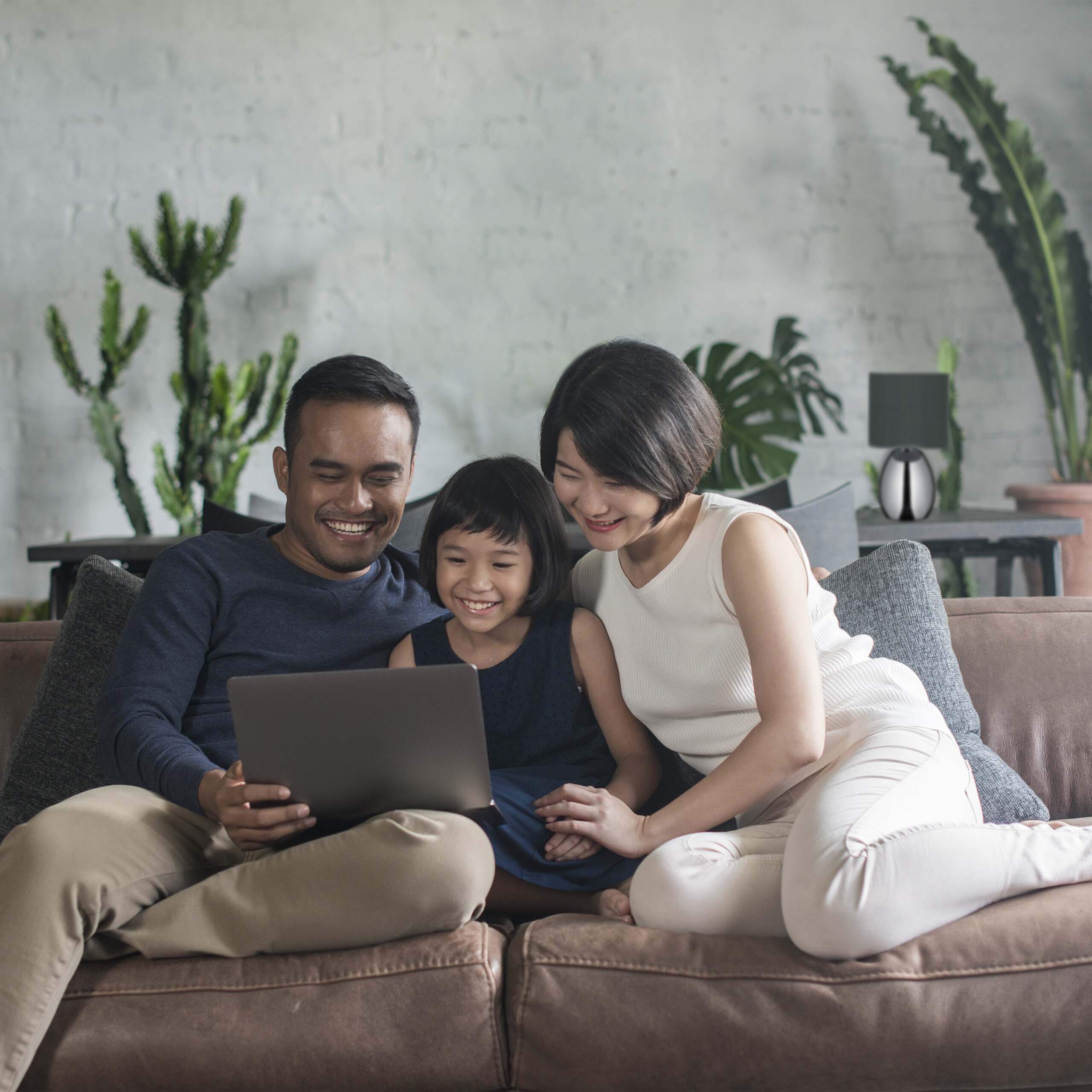 Eine lächelnde dreiköpfige Familie sitzt auf einem braunen Sofa und schaut in einem pflanzenreichen Wohnzimmer gemeinsam auf einen Laptop.