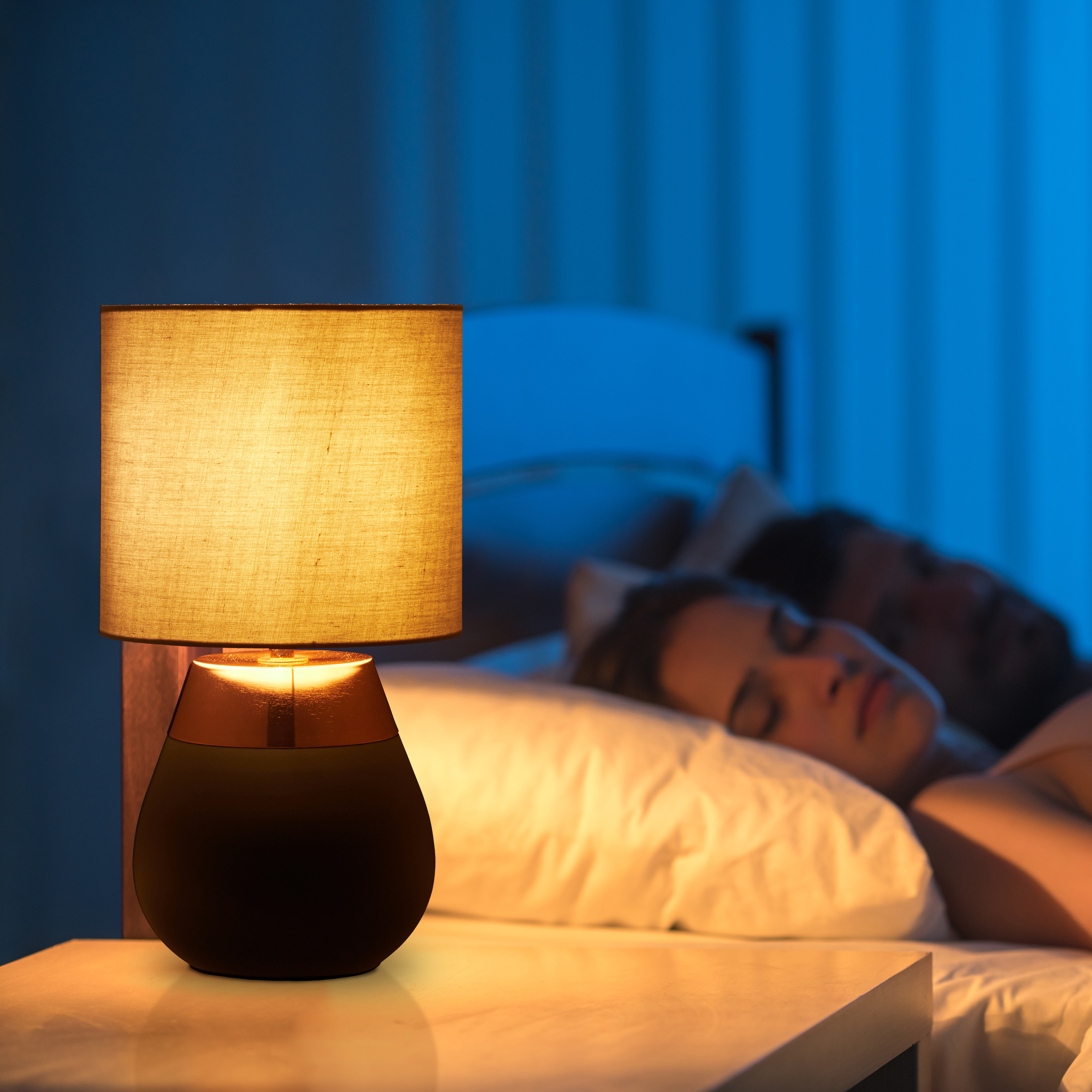 Eine brennende Nachttischlampe auf einem Nachttisch mit einem Paar im Hintergrund.