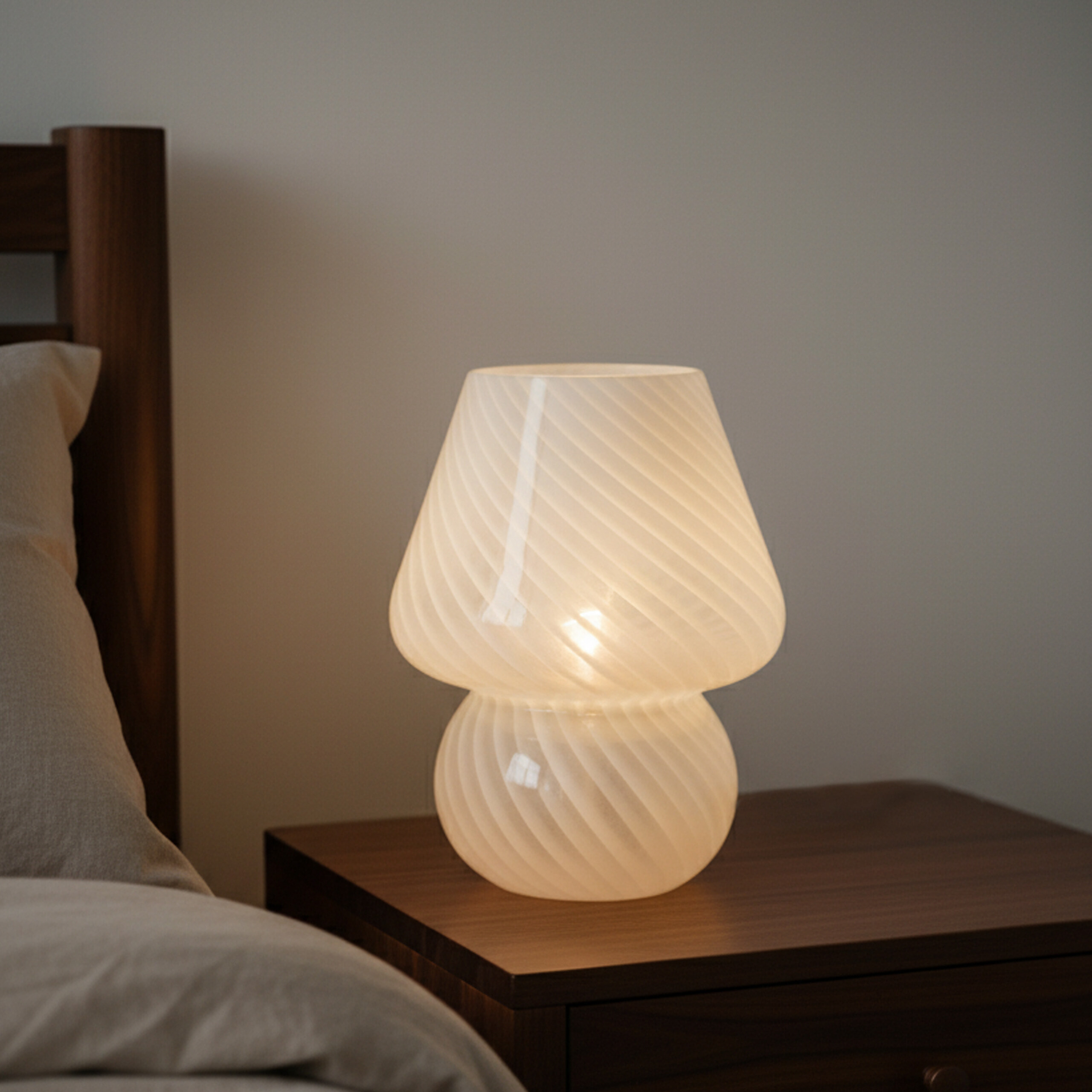 Eine weiße Glaslampe mit gedrehtem Muster steht auf einem Holznachttisch neben einem Bett.