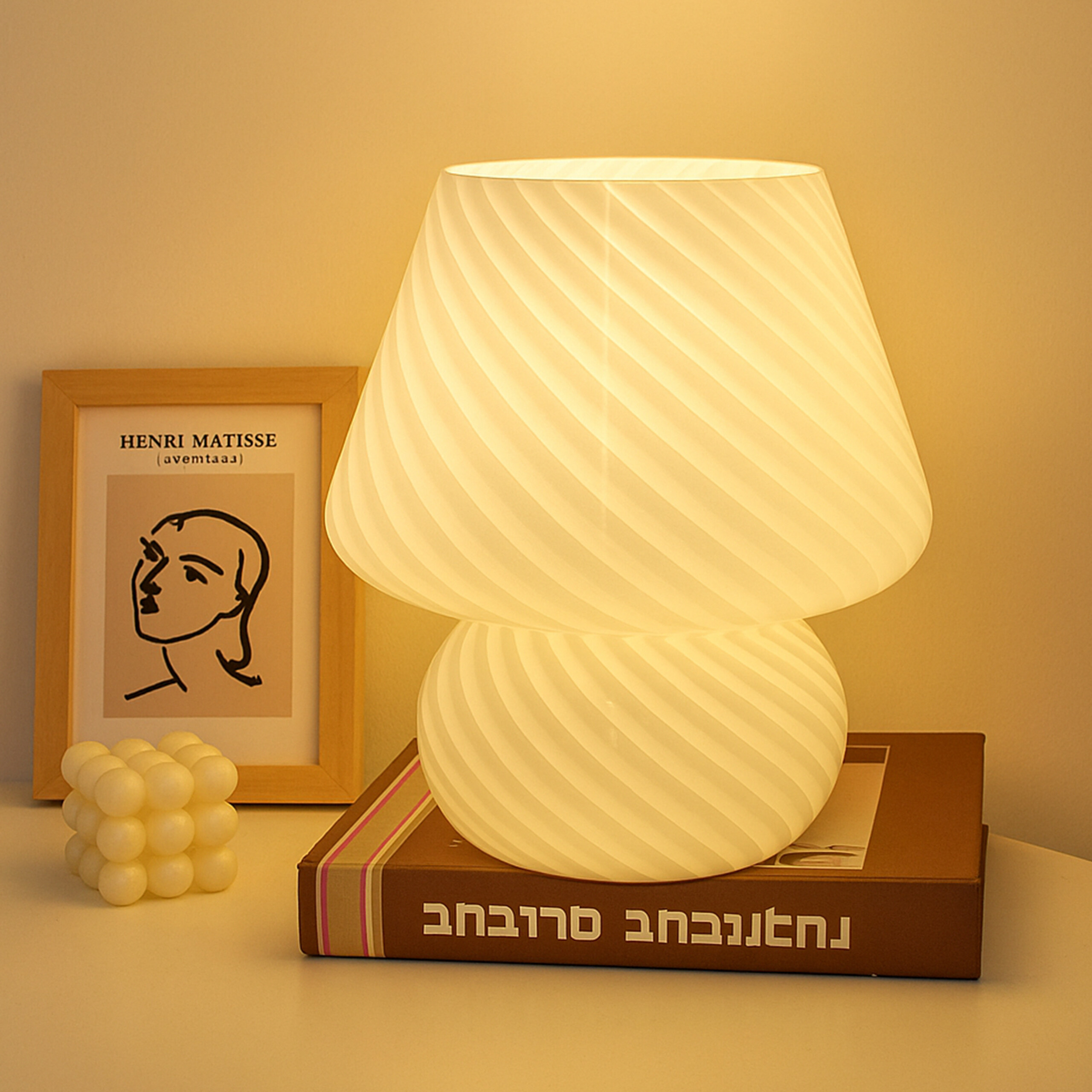 Eine leuchtende Tischlampe steht auf einem Buch, daneben ein kleiner weißer Block und ein gerahmtes Bild.