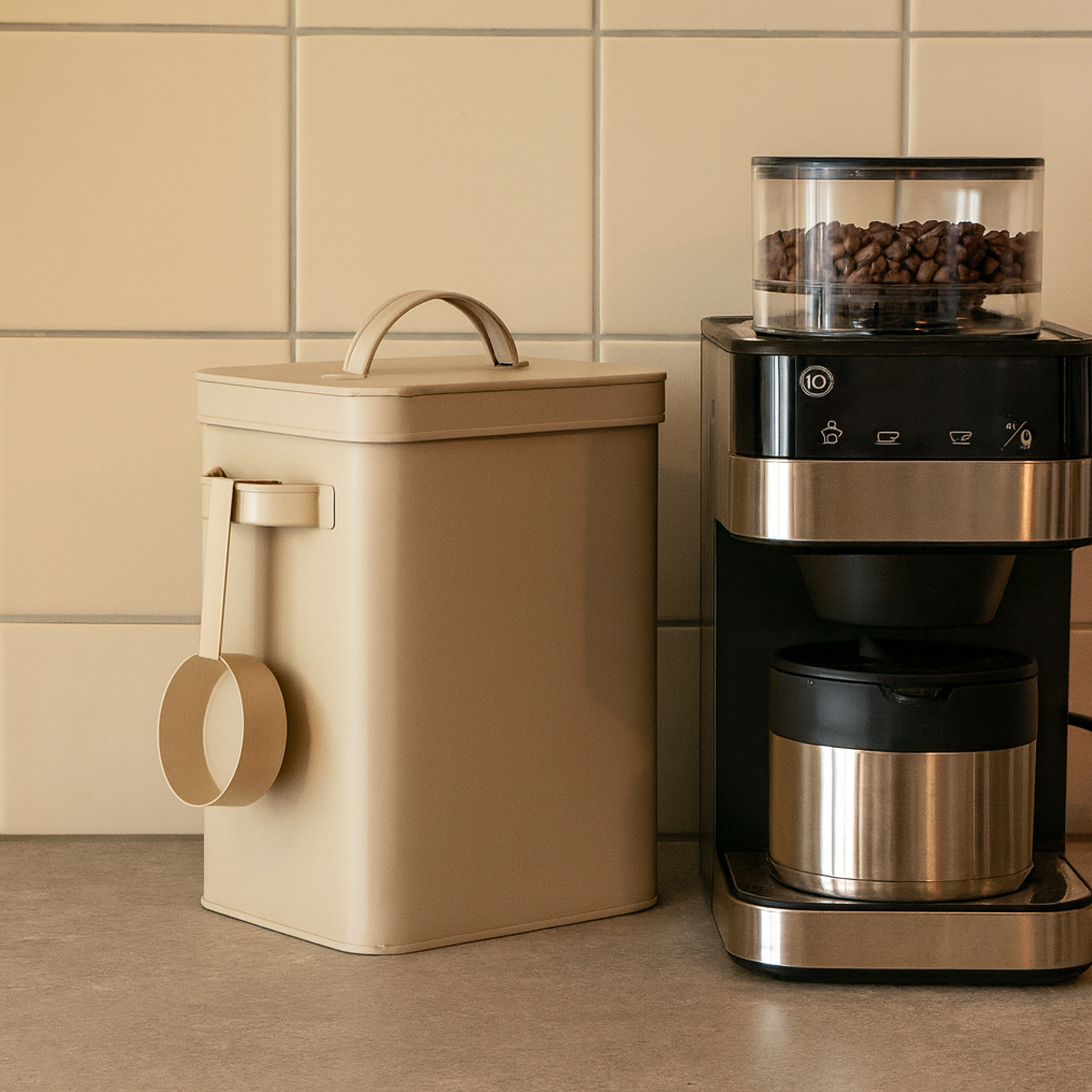 Beige Vorratsdose mit Griff und Löffel neben einer schwarz-silbernen Kaffeemaschine auf einer Küchenzeile.