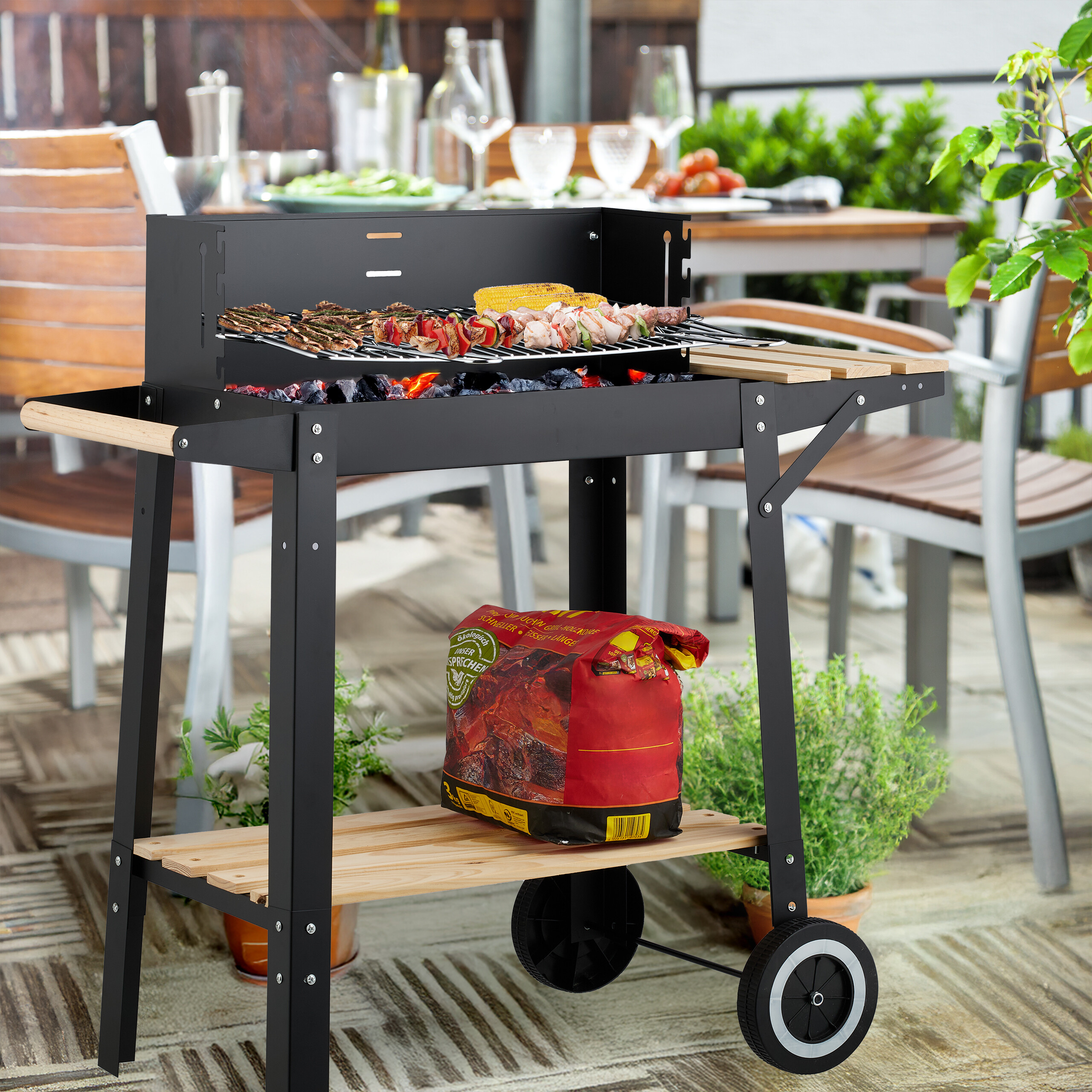 Ein schwarzer Holzkohlegrill mit glühender Kohle, grillendem Fleisch und Mais, auf einer Terrasse.