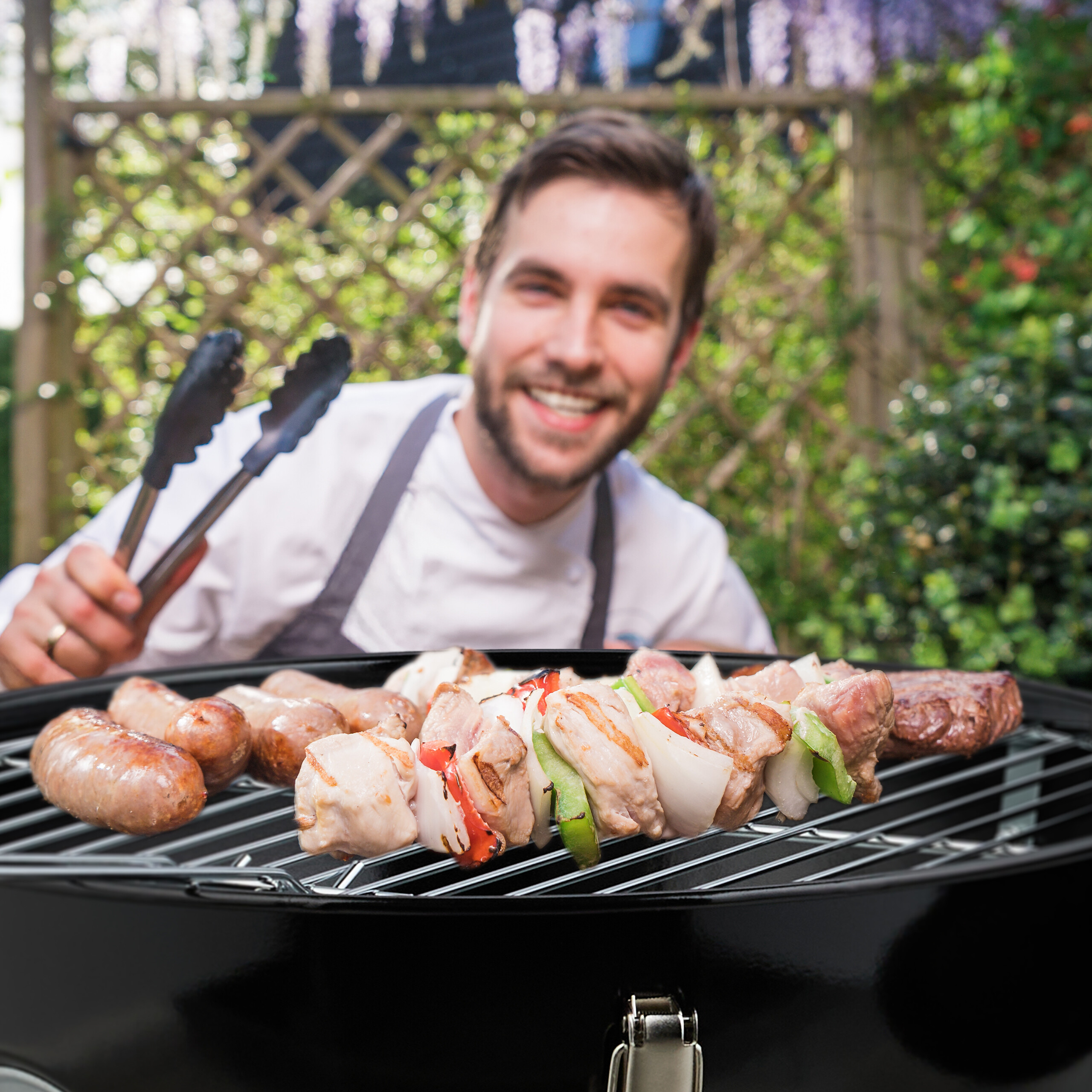 Ein lächelnder Koch grillt Würstchen und Fleischspieße im Garten.