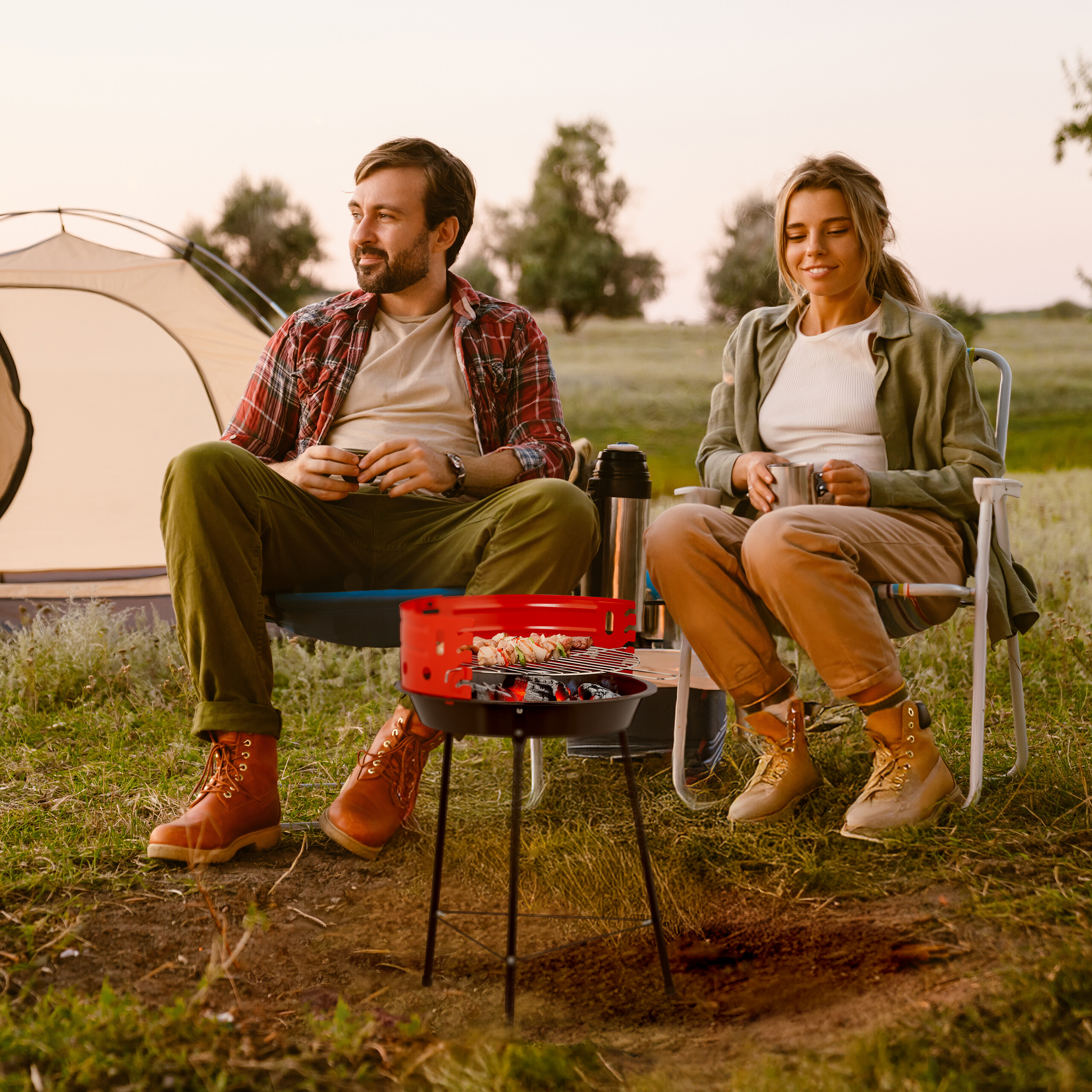 Ein Paar sitzt vor einem Zelt beim Camping, trinkt aus Tassen und kocht an einem kleinen Grill.