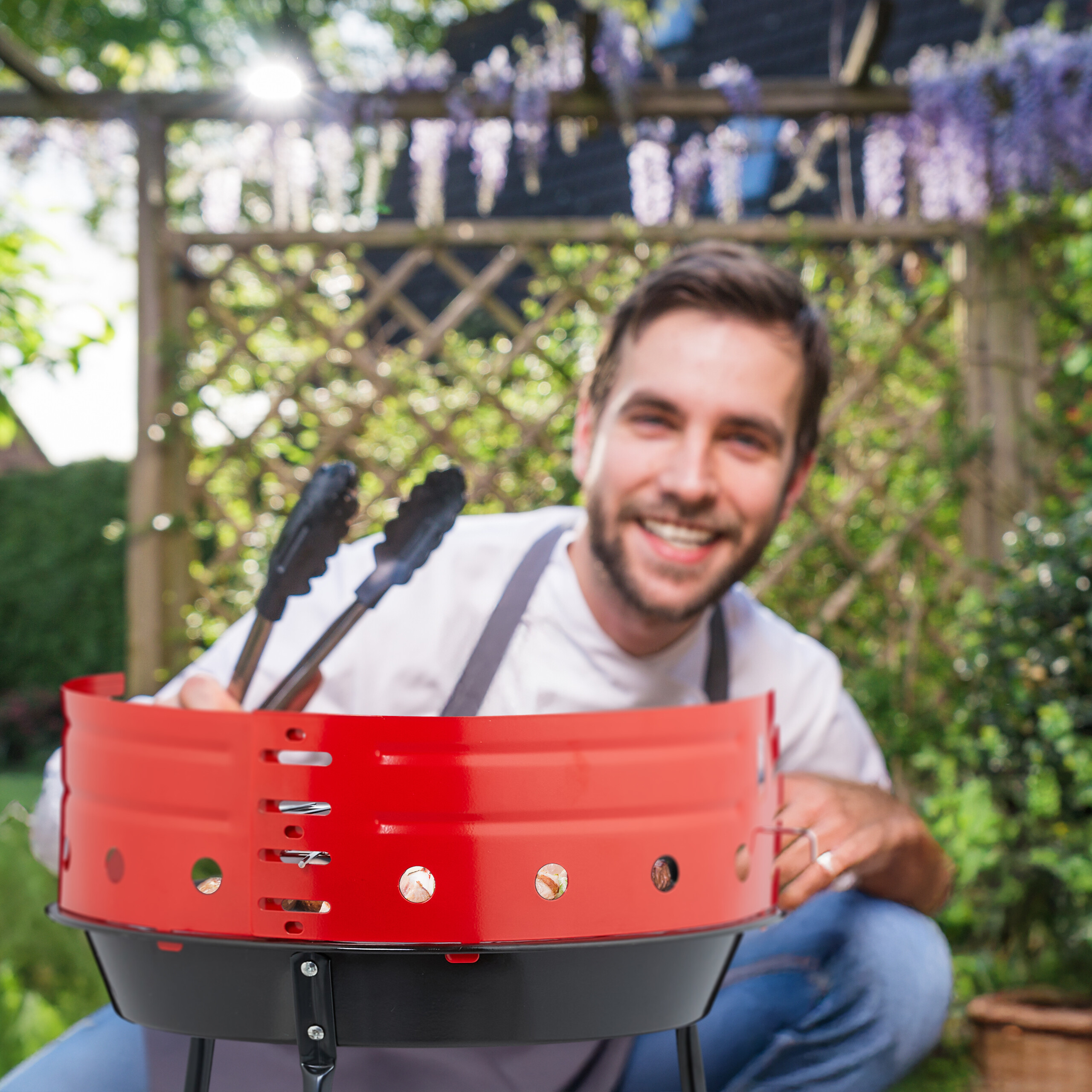 Ein lächelnder Mann in einer Kochschürze hält eine Grillzange über einem roten Holzkohlegrill in einem Garten.