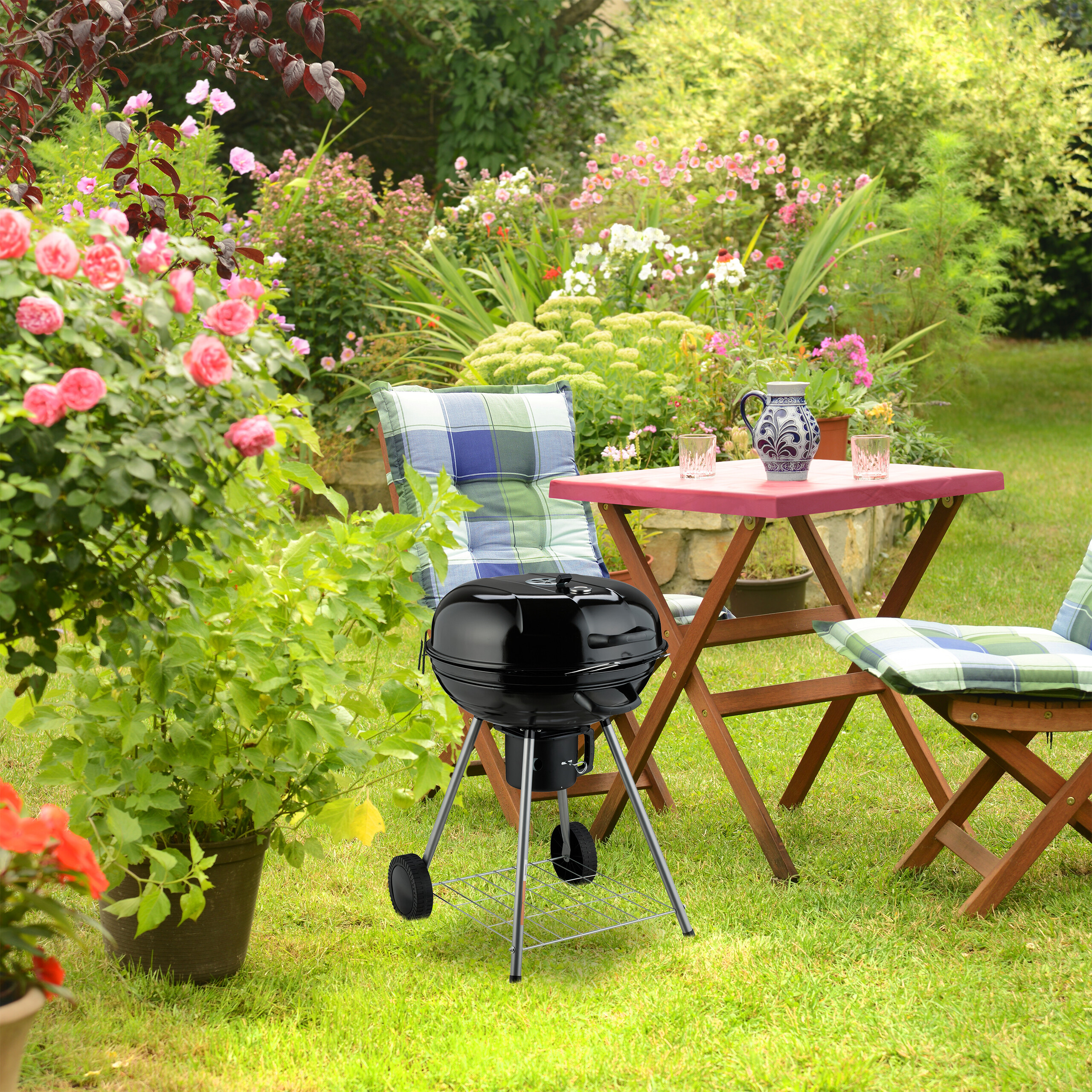 Ein schwarzer Kugelgrill und Holztische mit bunten Kissen stehen in einem blühenden Garten.