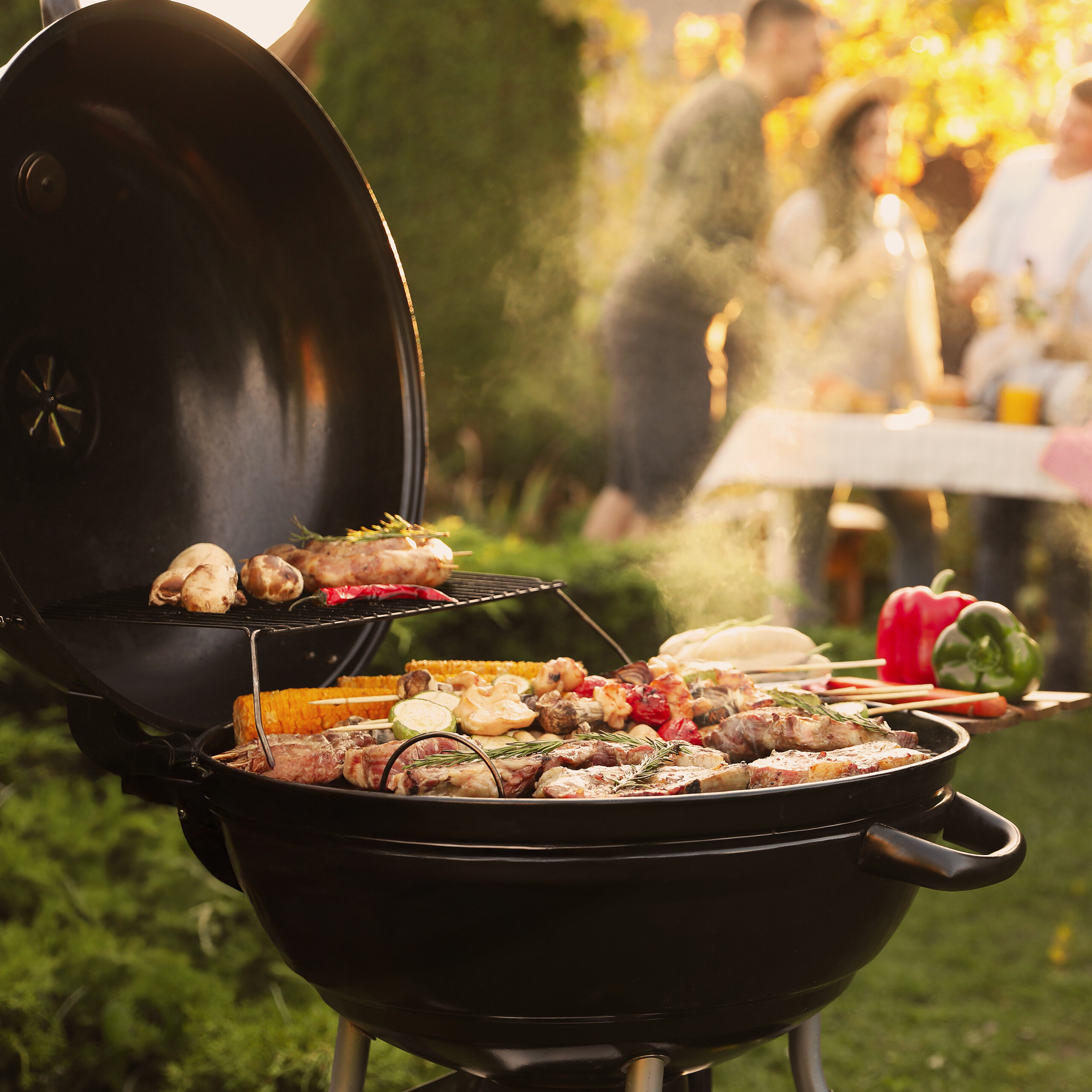 Fleisch, Pilze und Gemüse grillen auf einem runden Grill, während verschwommene Personen im Hintergrund feiern.