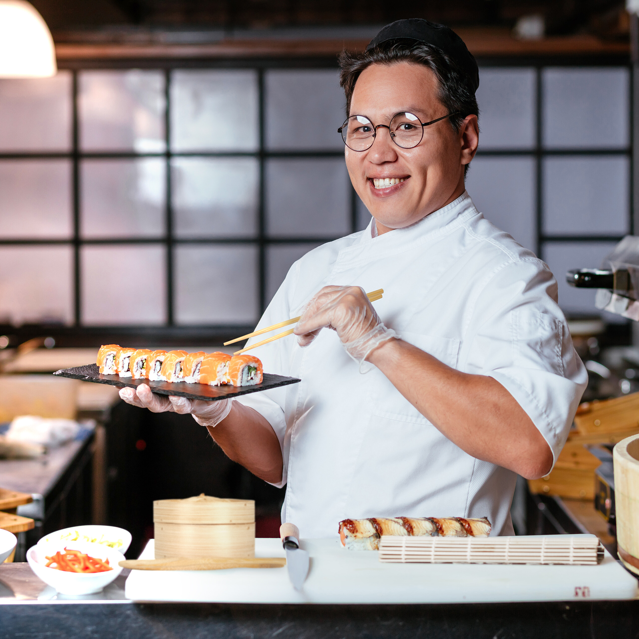 Ein lächelnder asiatischer Koch in weißer Uniform und Brille hält einen Teller Sushi in einer Hand und Stäbchen in der anderen.