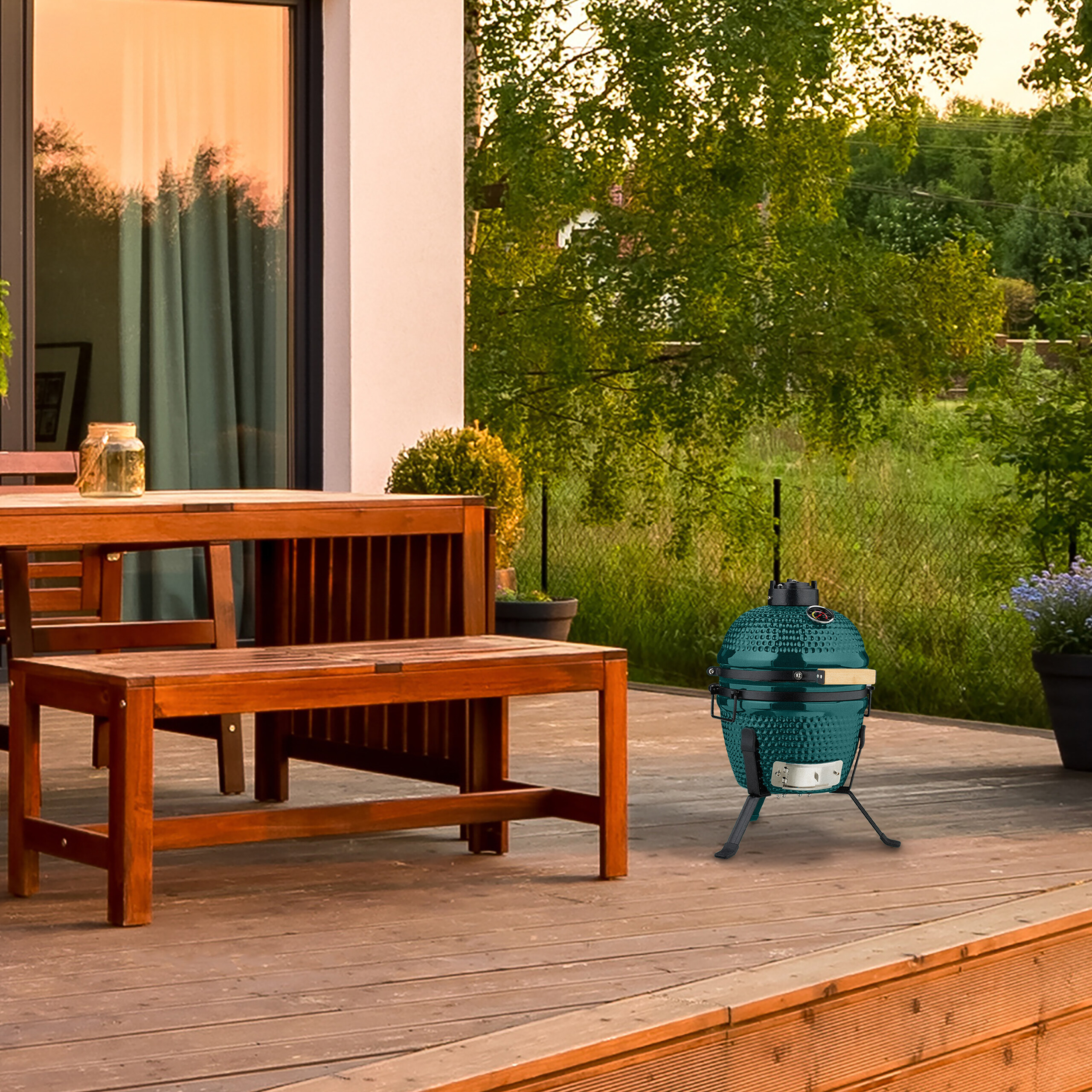 Grüner Kamado-Grill auf einer Holzterrasse vor einem Haus mit Tisch und Bank.