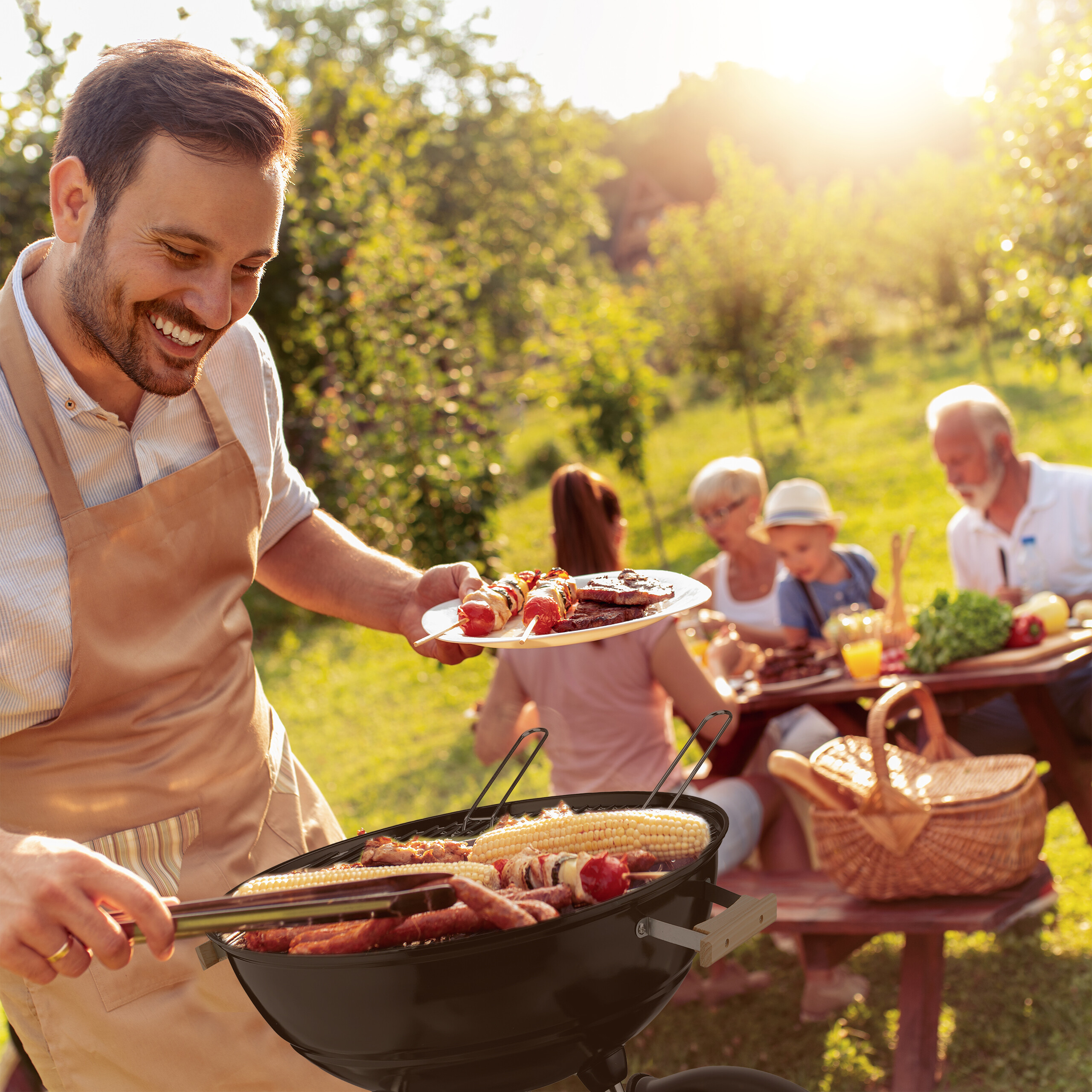 Mann in Schürze grillt Fleisch und Maiskolben im Garten, während Familie im Hintergrund am Tisch sitzt.