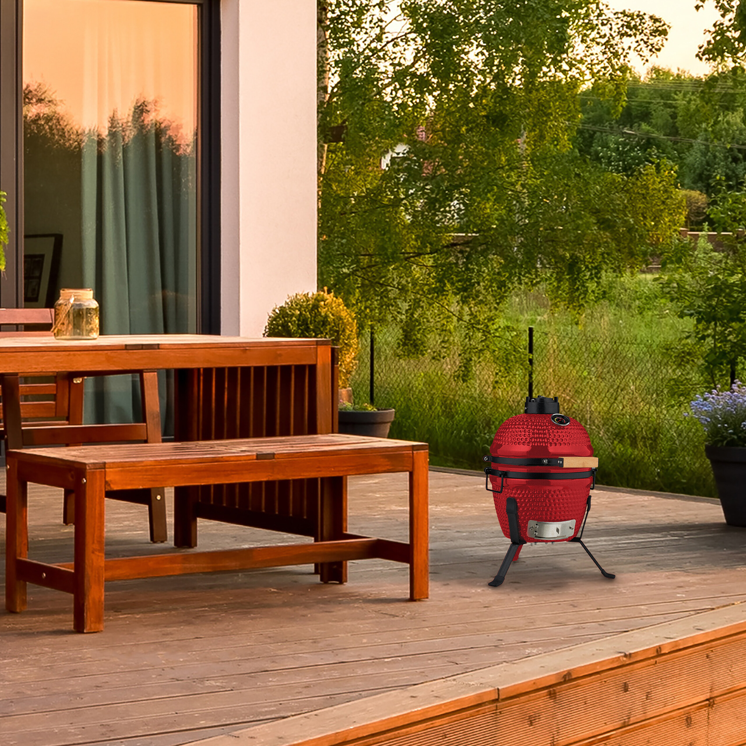 Ein roter Kamado-Grill steht auf einer Holzterrasse neben einem Holztisch mit Bank, im Hintergrund Bäume.