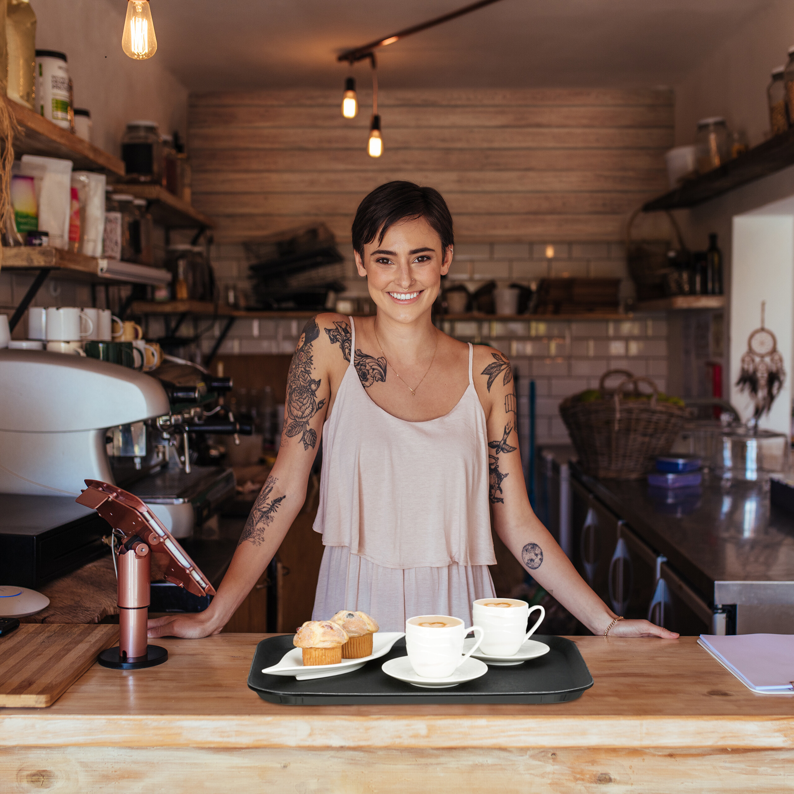 Eine lächelnde tätowierte Barista steht hinter einem Holztresen mit Kaffee und Muffins.