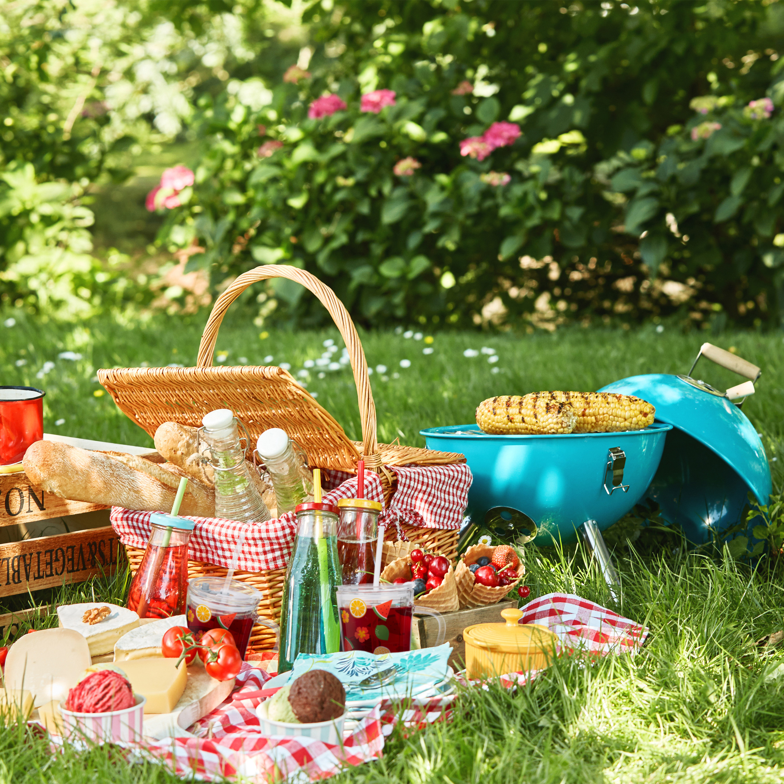 Ein sonniges Picknick im Park mit Weidenkorb, Getränken, Käse, Beeren und blauem Grill mit Mais.