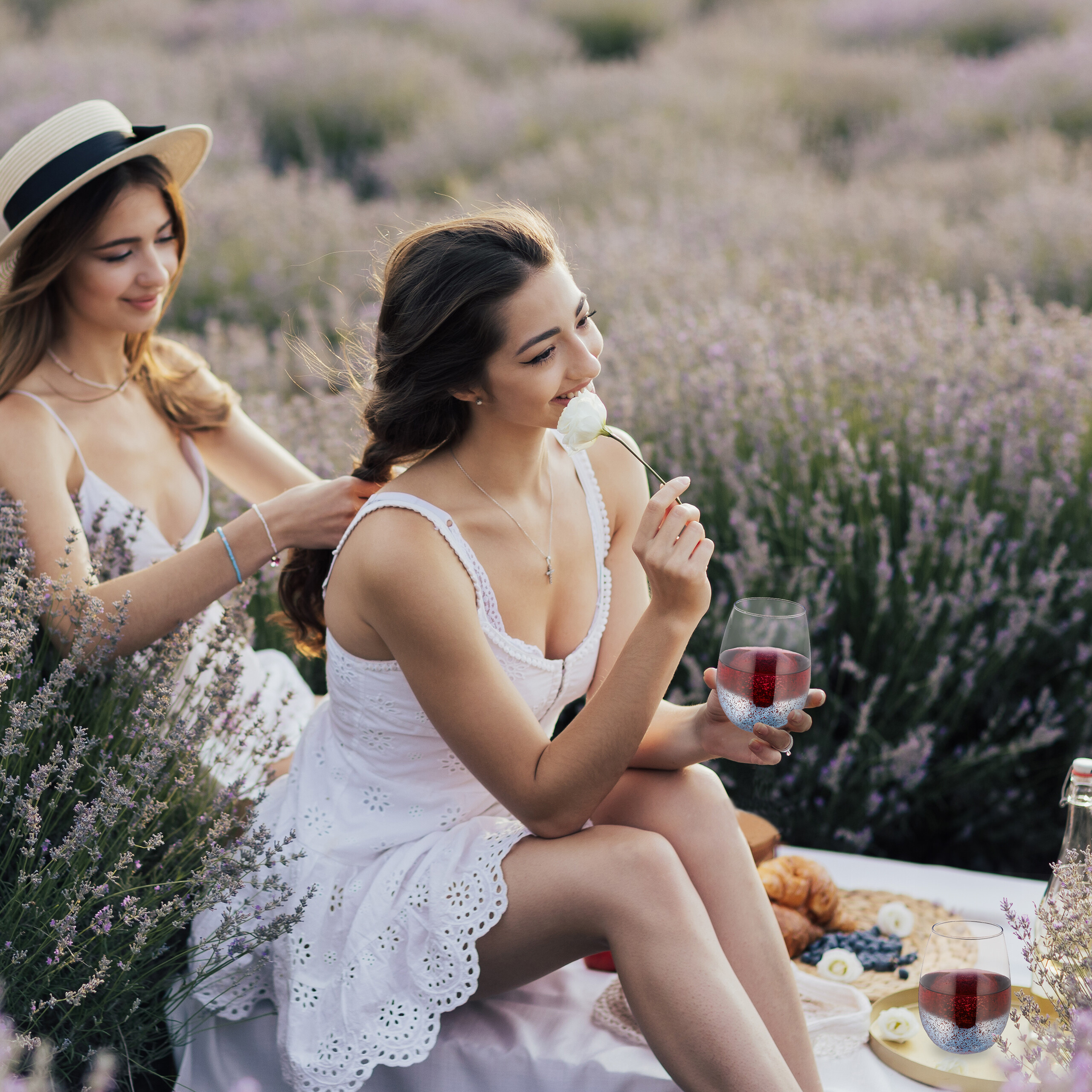 Zwei Frauen in weißen Kleidern in einem Lavendelfeld, eine flechtet Haare, während die andere eine Blume und Wein hält.
