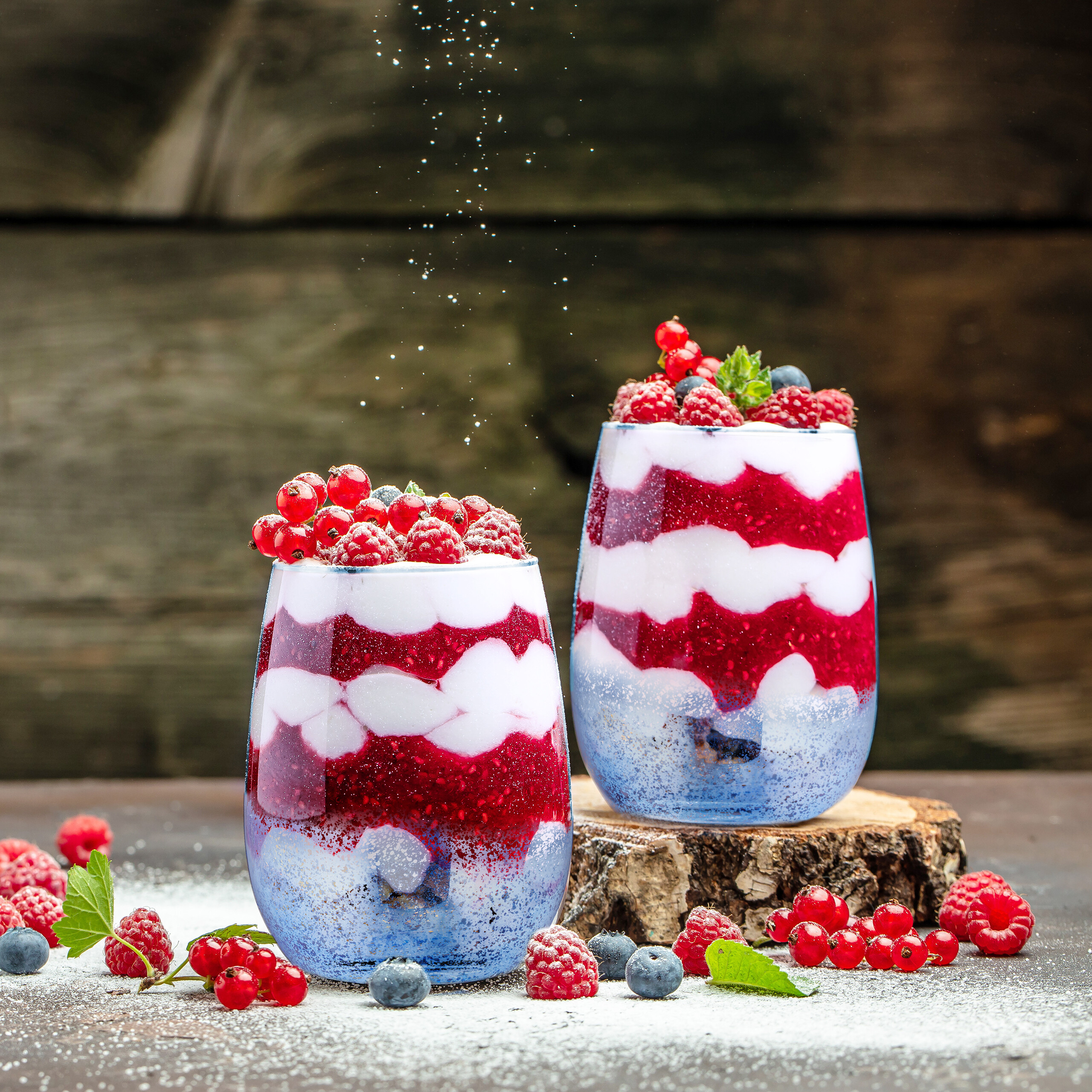 Zwei Gläser Beicht-Schichtdessert mit Joghurt und rotem Fruchtpüree, gekrönt mit frischen Beeren und Zucker.