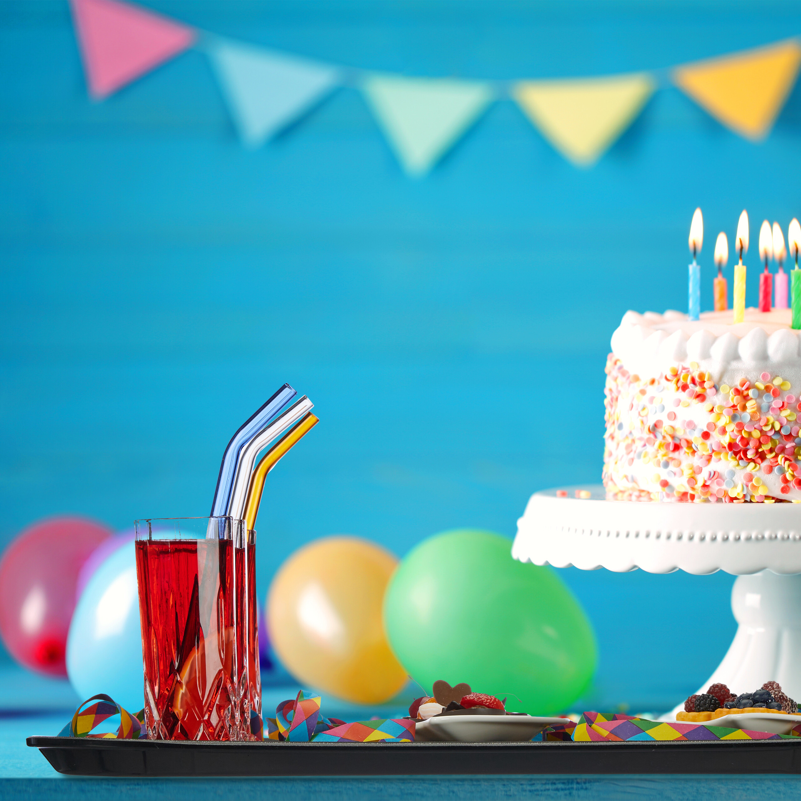 Eine Geburtstagstorte mit Kerzen, ein Getränk mit Strohhalmen und bunte Luftballons vor einer blauen Wand.