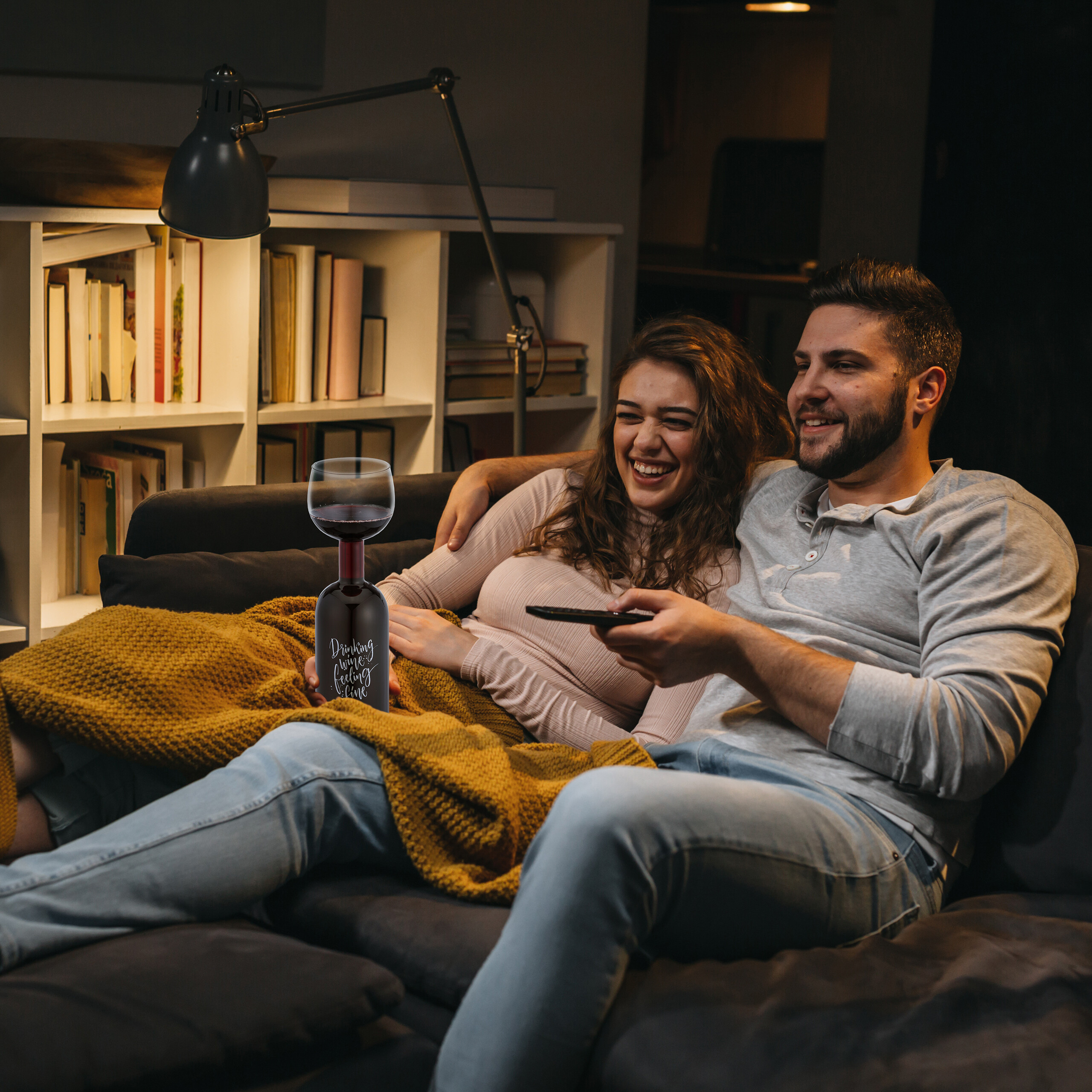 Ein glückliches Paar sitzt nachts auf einem Sofa und lacht beim Fernsehen, gedeckt mit einer Decke.
