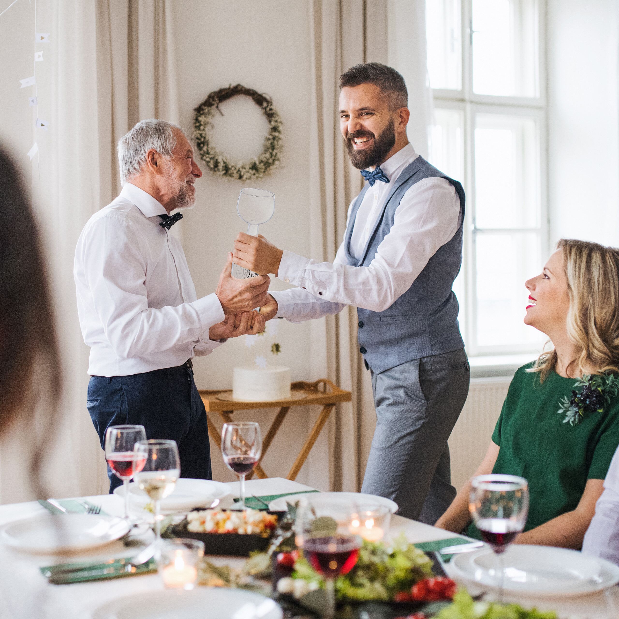 Ein jüngerer Mann überreicht einem älteren Mann ein großes Glas bei einem Familienessen.