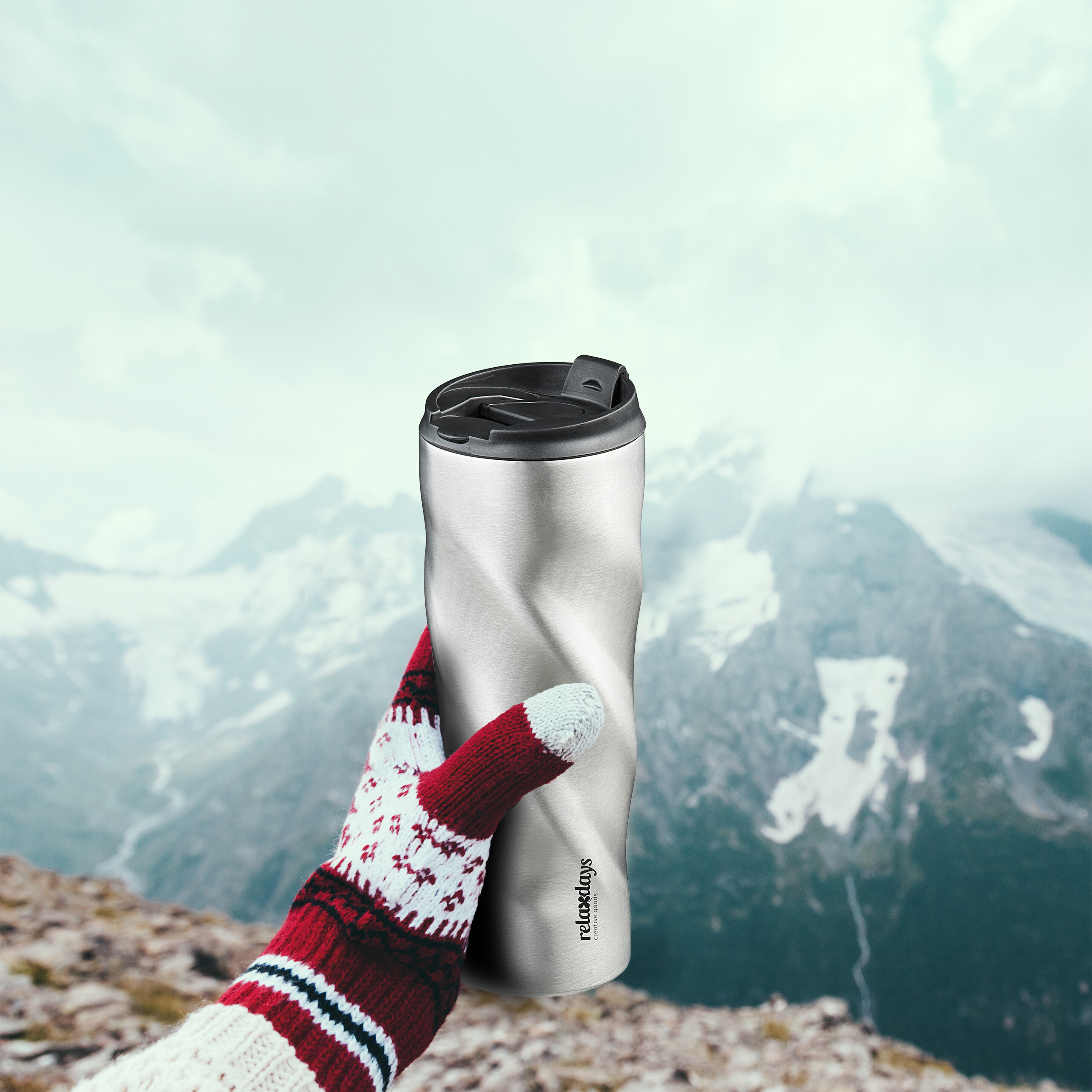 Eine Hand in einem rot-weißen Strickhandschuh hält einen Edelstahlbecher vor einer schneebedeckten Bergkulisse.