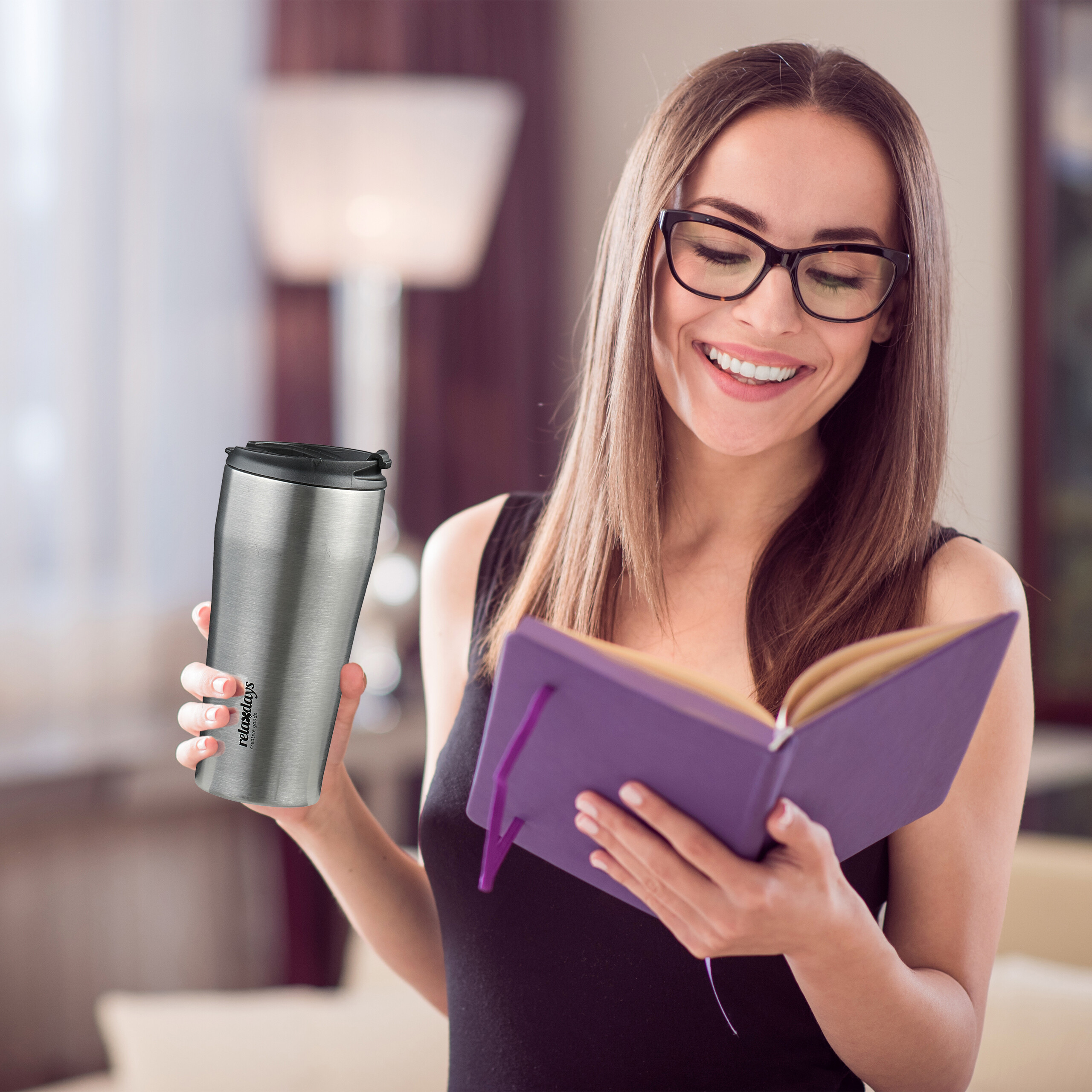 Lchelnde Frau mit Brille hlt einen silbernen Reisebecher und liest ein lila Notizbuch.