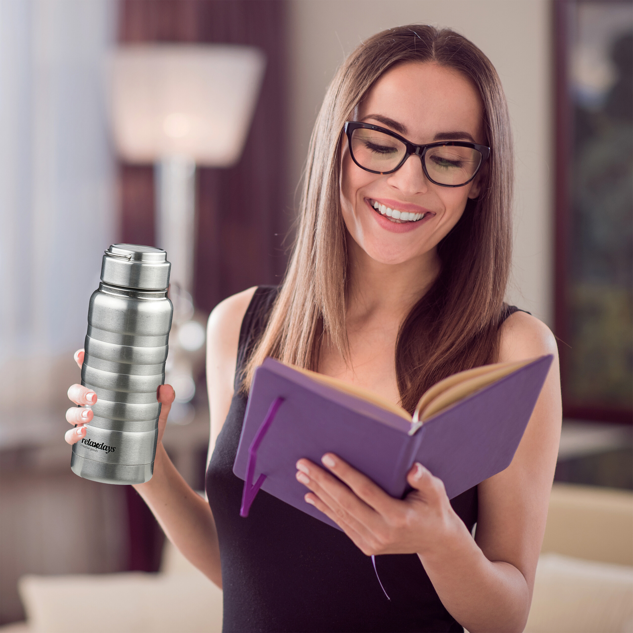 Eine lächelnde Frau mit Brille liest in einem lilafarbenen Notizbuch und hält eine Edelstahl-Trinkflasche.