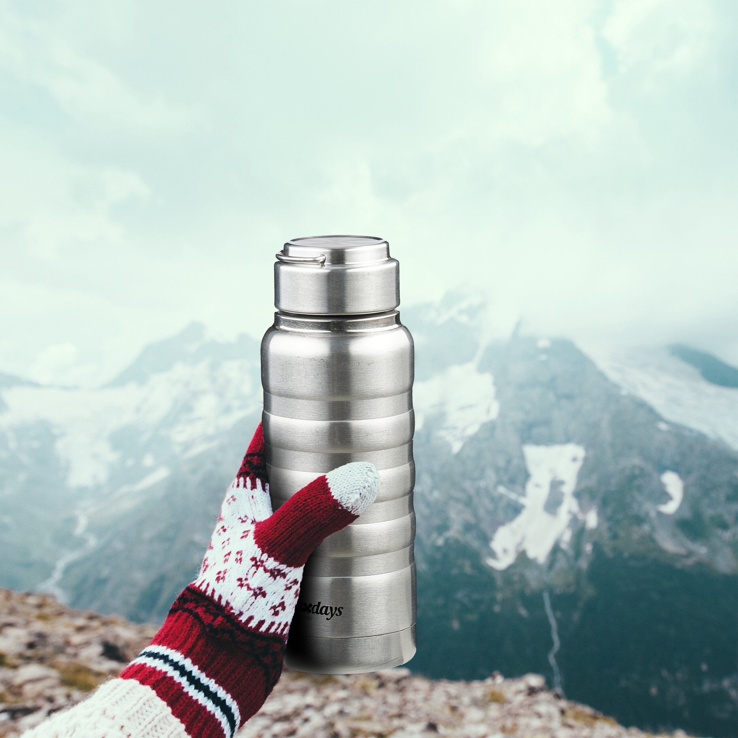Eine behandschuhte Hand hält eine silberne Thermoskanne vor verschneiter Bergkulisse.