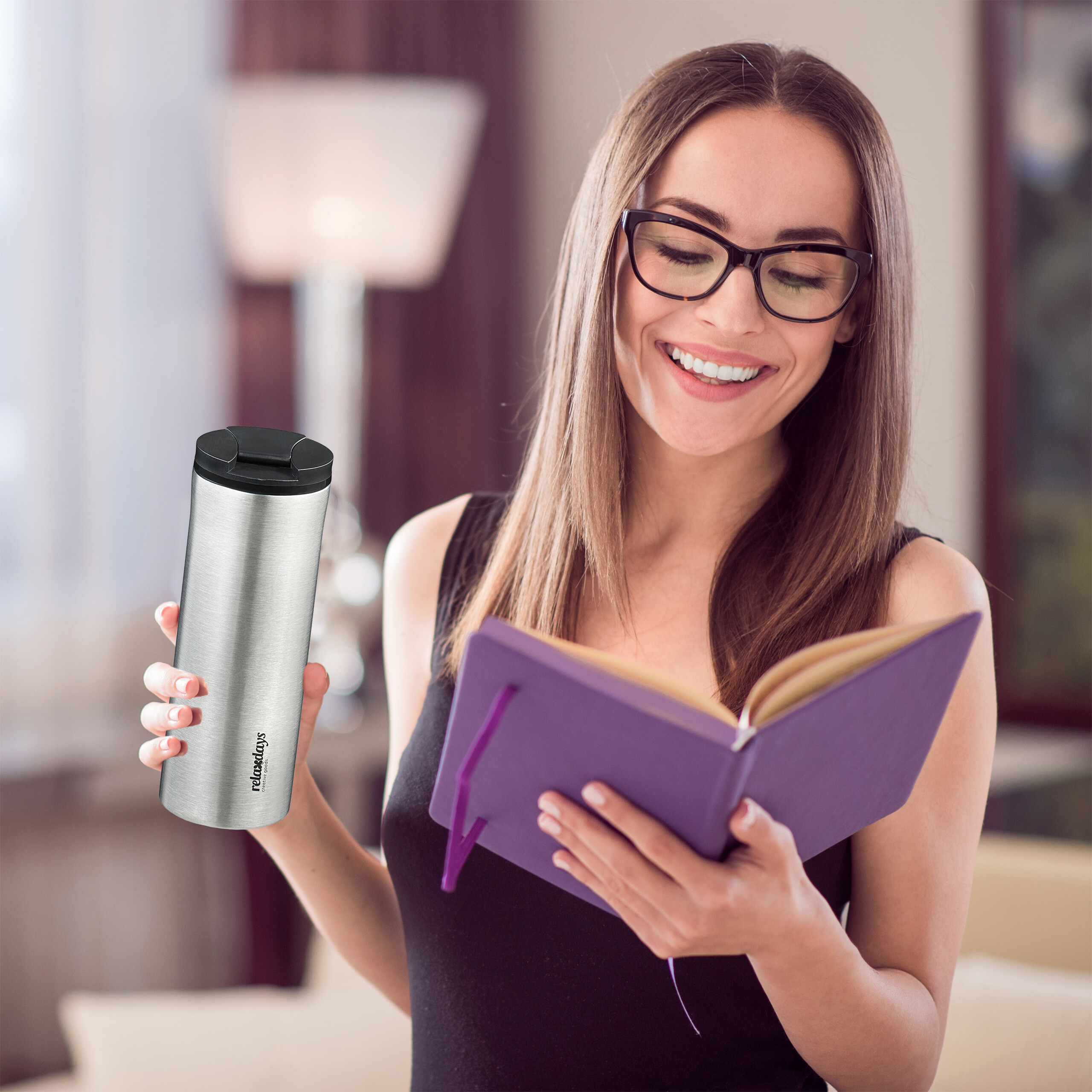 Eine lächelnde Frau mit Brille liest ein lilafarbenes Notizbuch und hält einen silbernen Thermobecher.