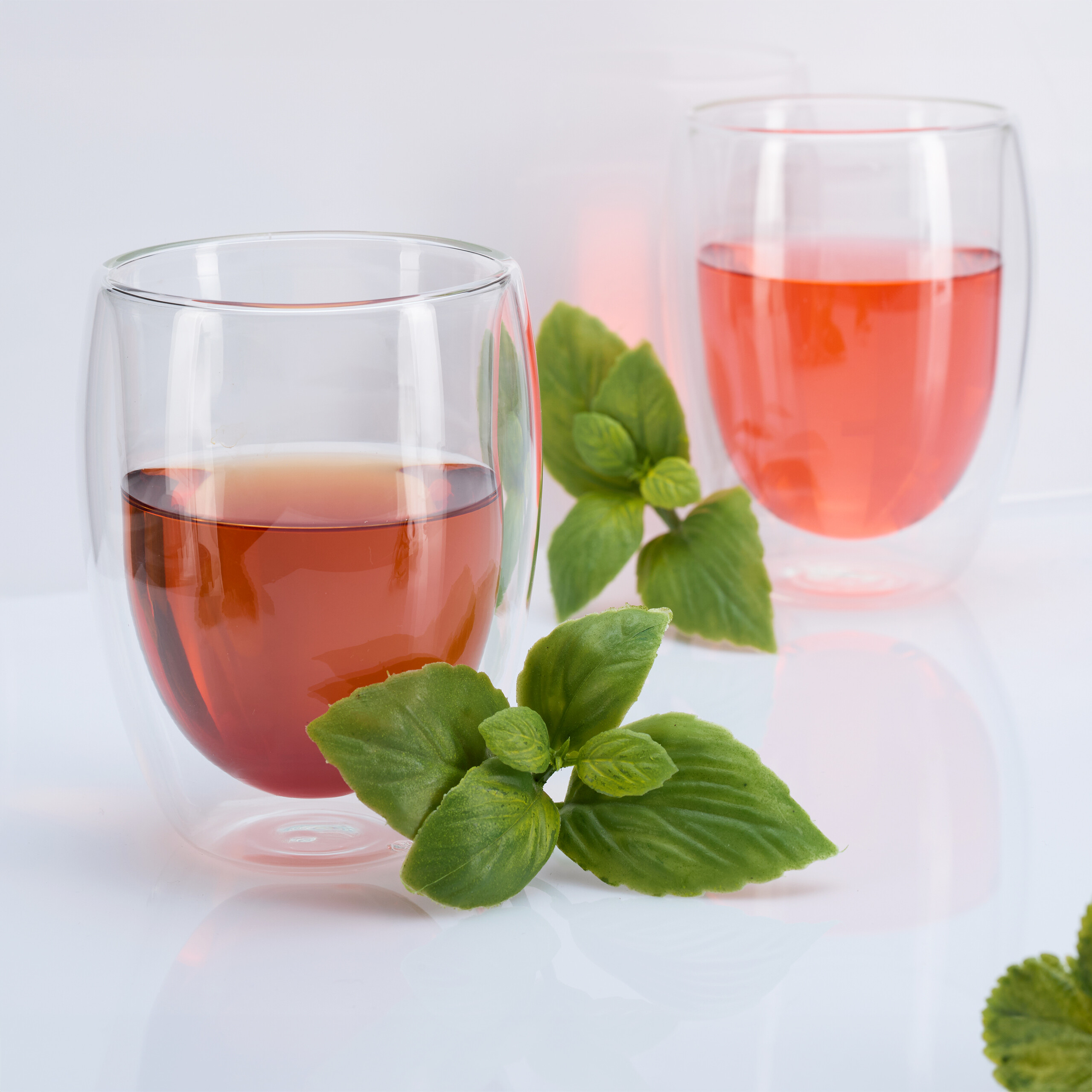Zwei doppelwandige Gläser mit rosa Getränk und grünen Minzblättern auf weißer Fläche.