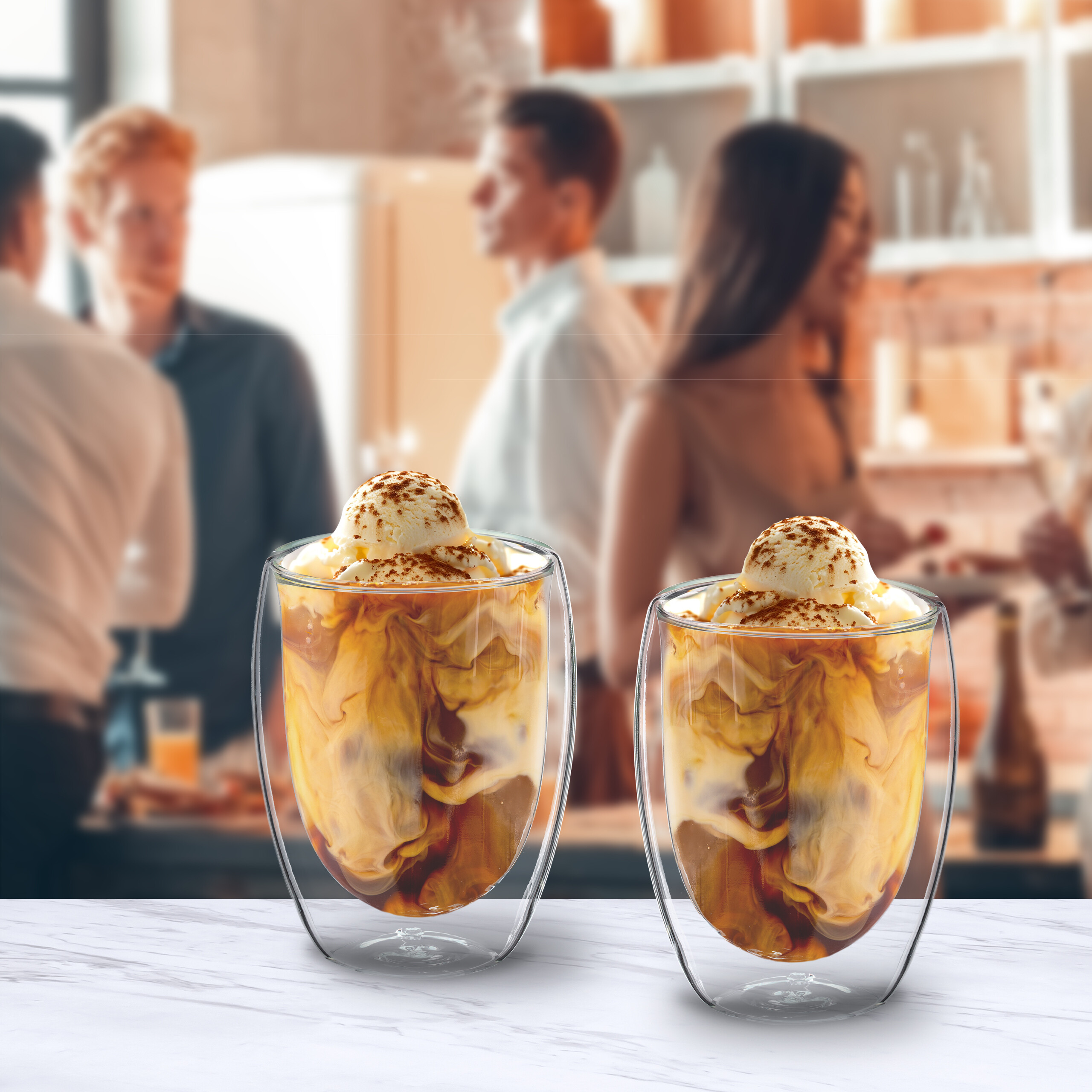 Zwei Eiskaffee-Gläser mit Vanilleeis stehen auf einer weißen Theke; im Hintergrund unterhalten sich Personen.