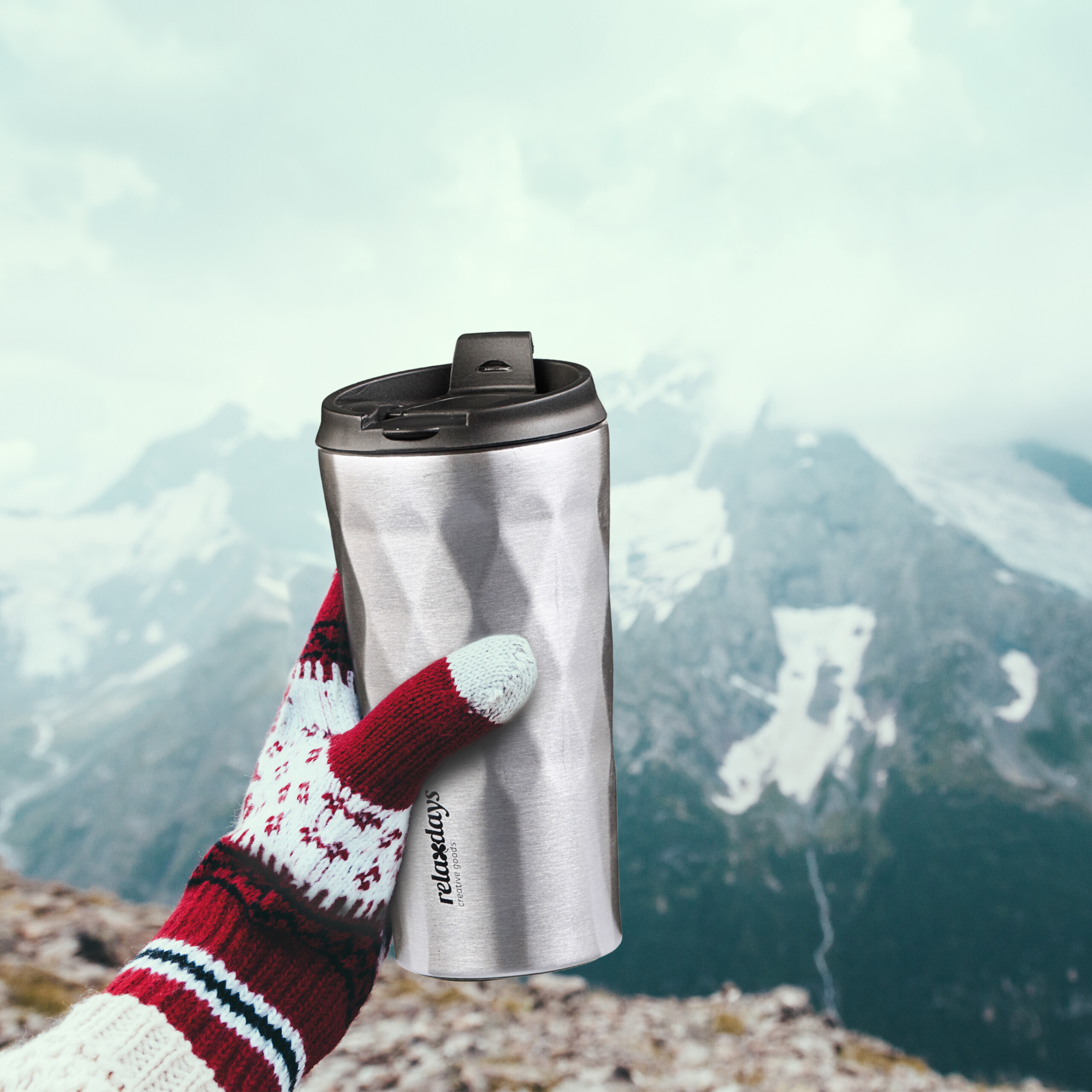 Hand im gemusterten Handschuh hält Thermobecher vor verschneiter Bergkulisse.
