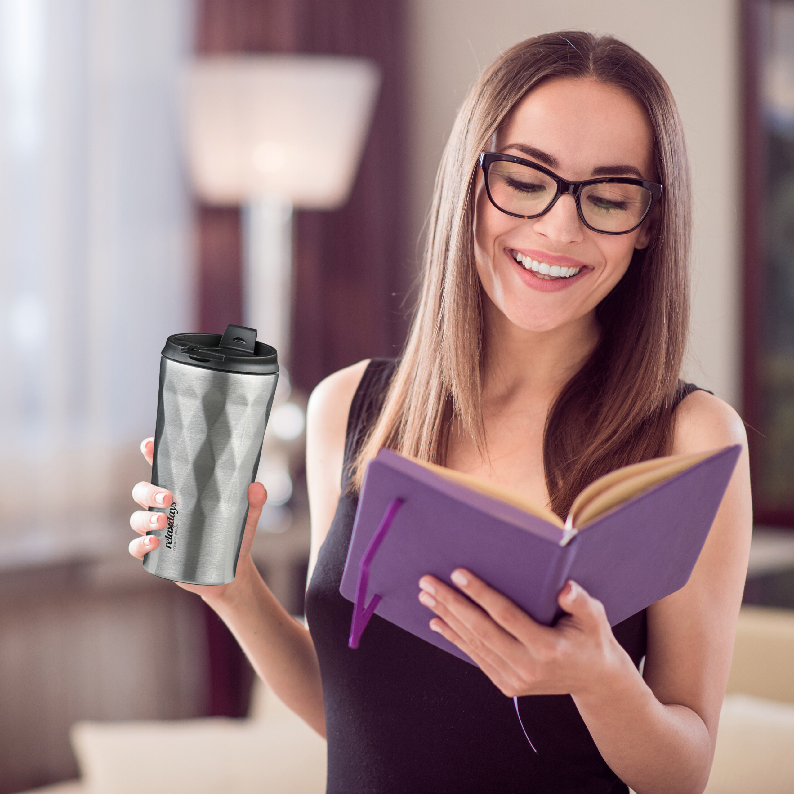 Eine lächelnde Frau mit Brille liest ein lilafarbenes Notizbuch und hält einen silbernen Thermobecher.