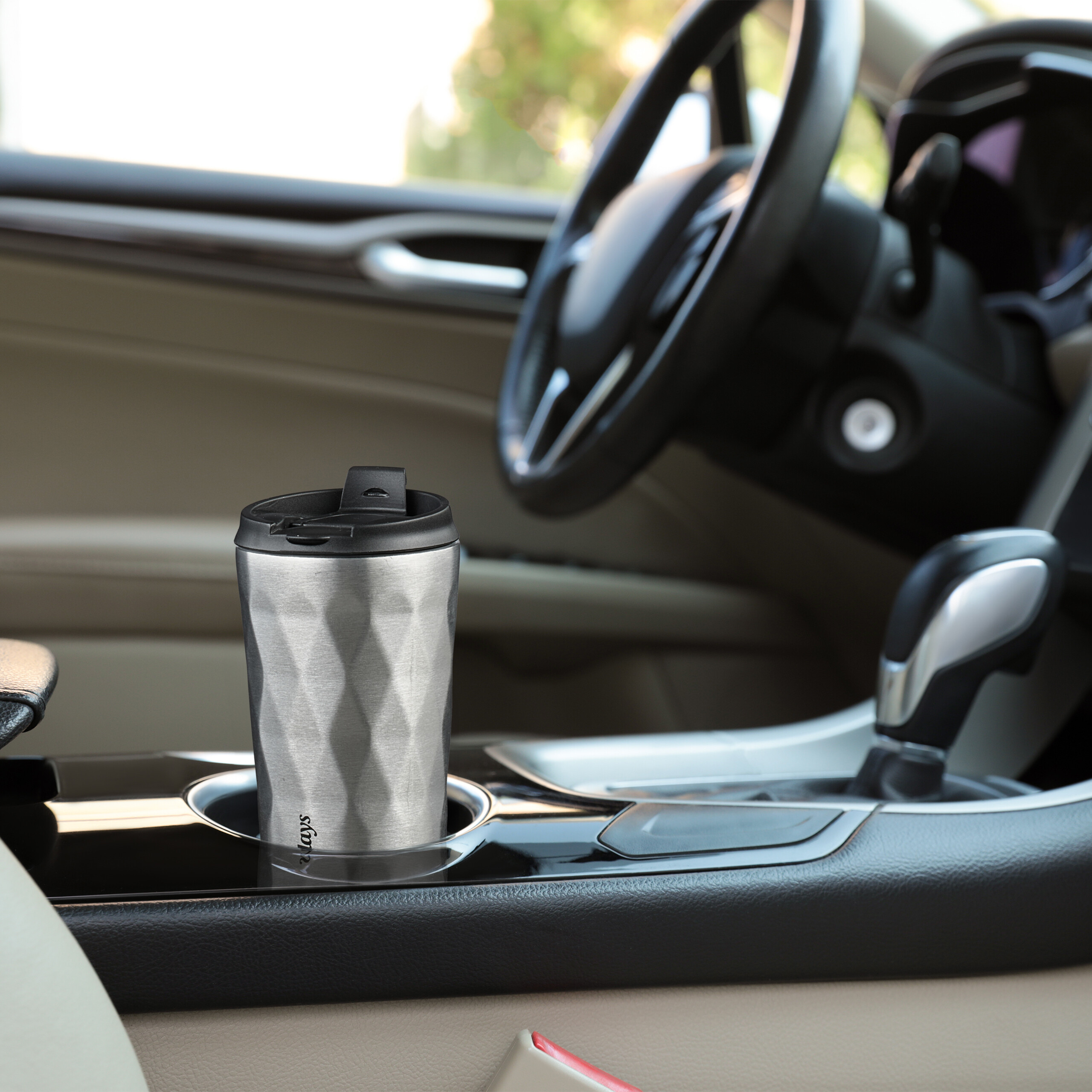 Ein silberner Thermobecher steht im Becherhalter der Mittelkonsole eines Autos, das Lenkrad ist im Hintergrund unscharf.