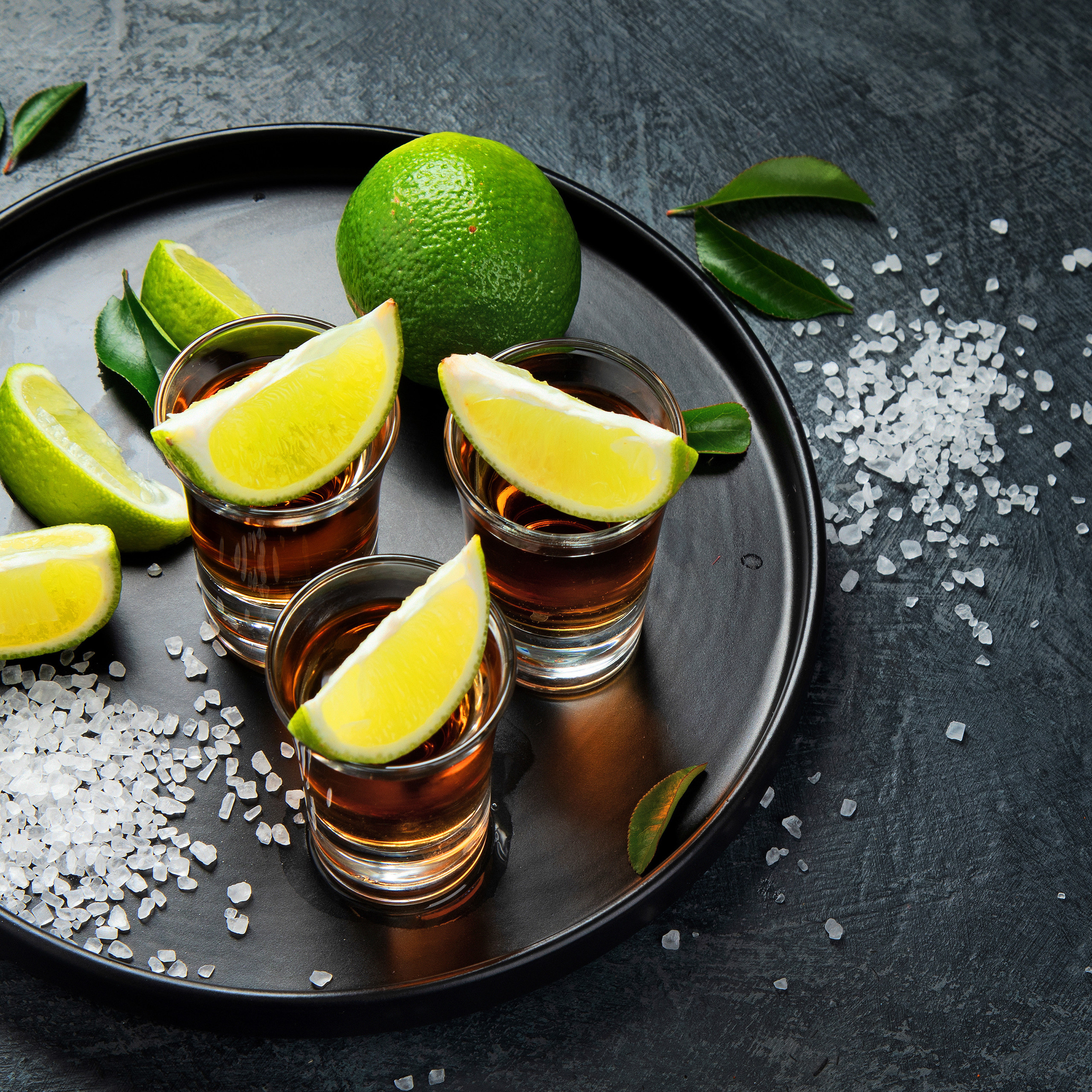 Drei Tequila-Shots mit Limettenscheiben und Salz auf schwarzem Tablett.