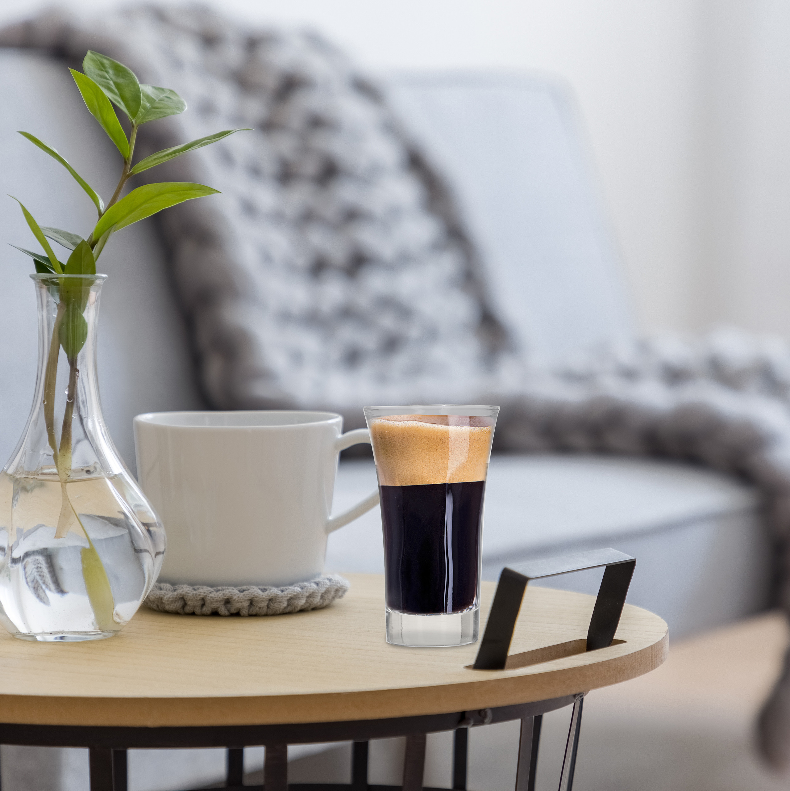Ein hölzerner Couchtisch mit einem Glas Kaffee, einer weißen Tasse und einer Vase mit grünen Zweigen.