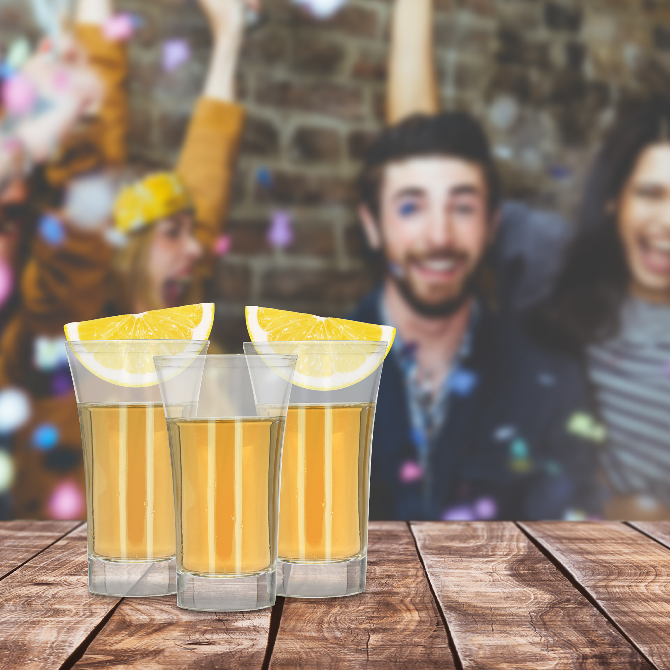 Drei Tequila-Shots mit Limettenscheiben stehen auf einem Holztisch, im Hintergrund feiern jubelnde Personen.