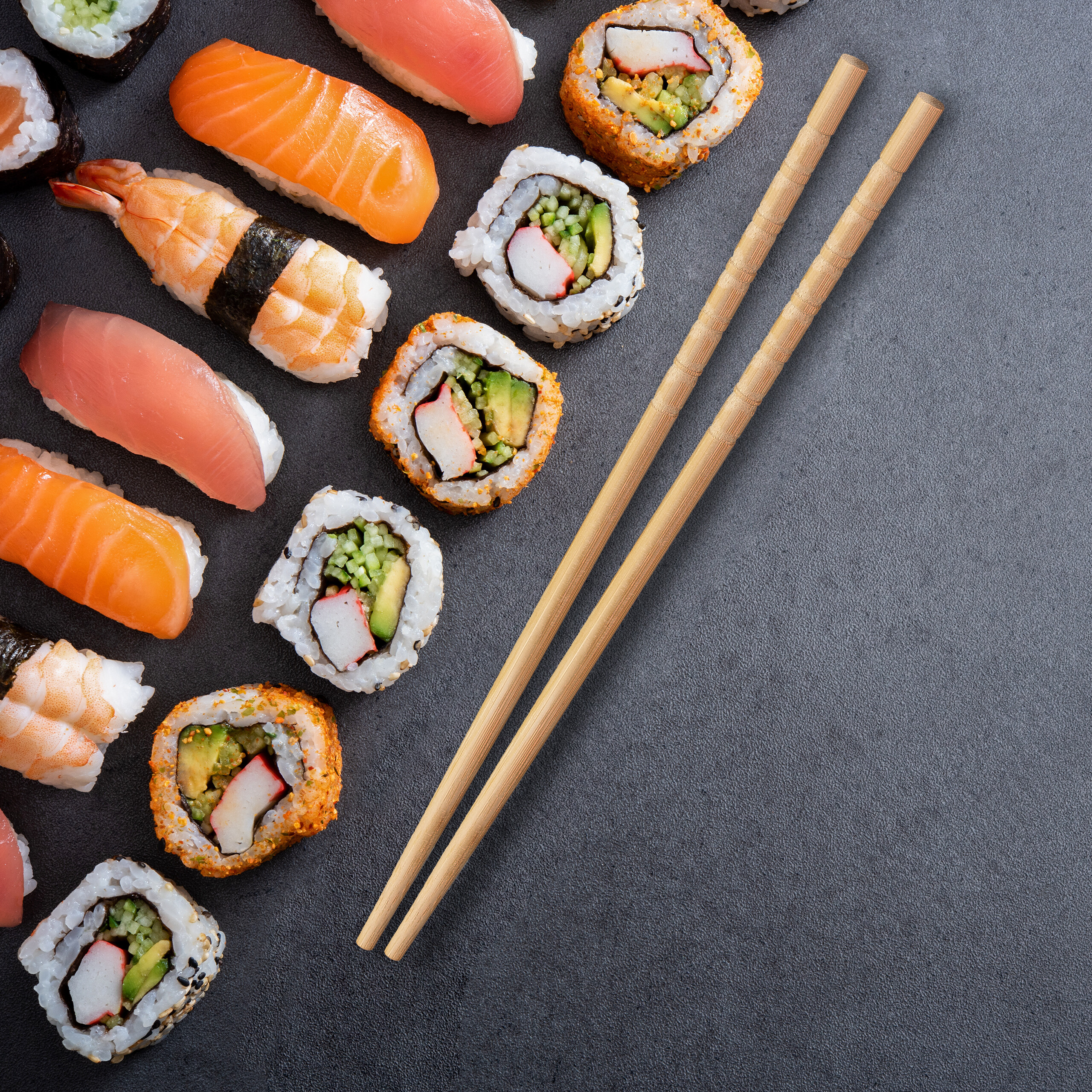 Eine Draufsicht auf verschiedene Sushi-Rollen und Nigiri auf dunkler Oberfläche mit Essstäbchen.