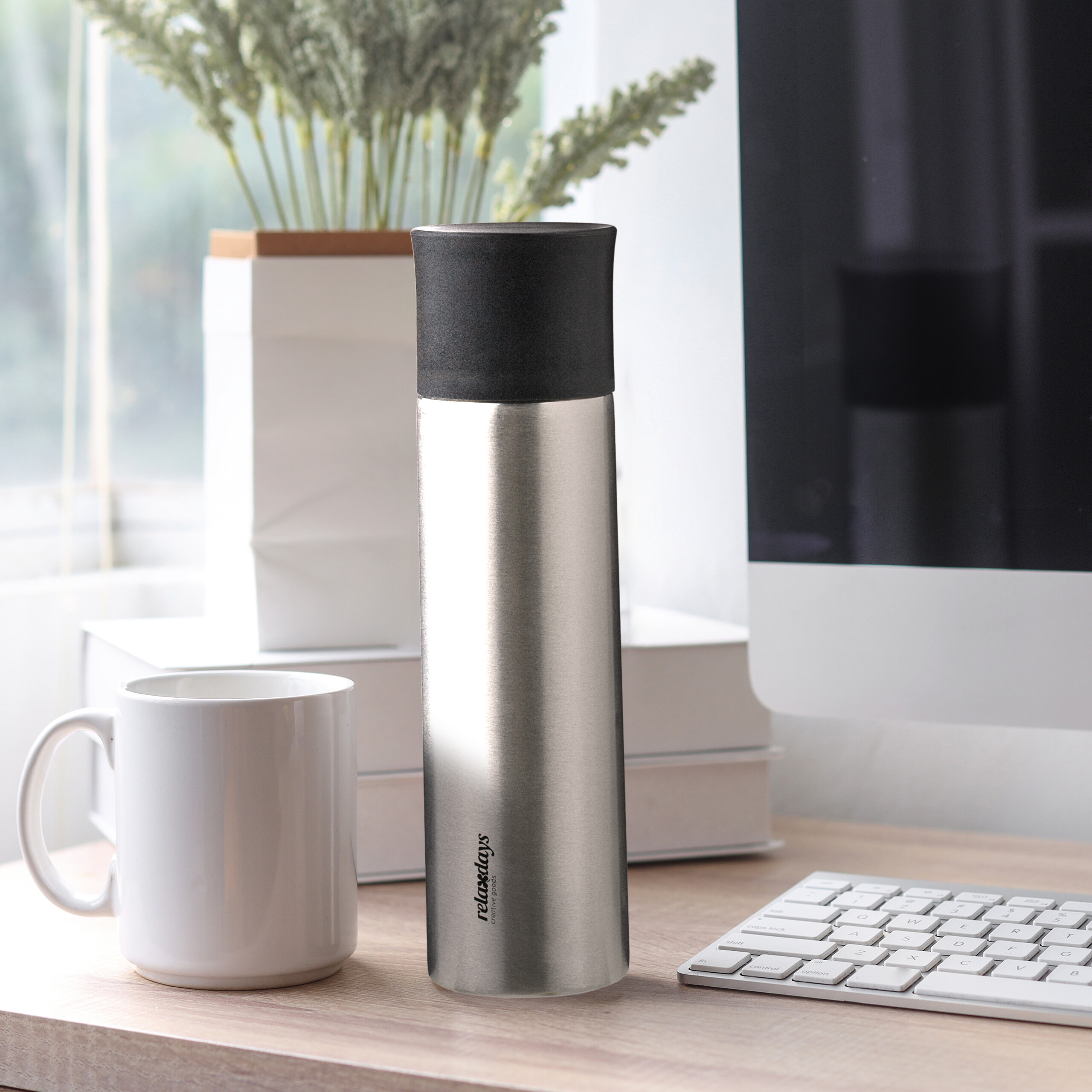 Eine Thermoskanne, weiße Tasse, Bücher, Pflanze, Tastatur und Monitor auf einem Schreibtisch.