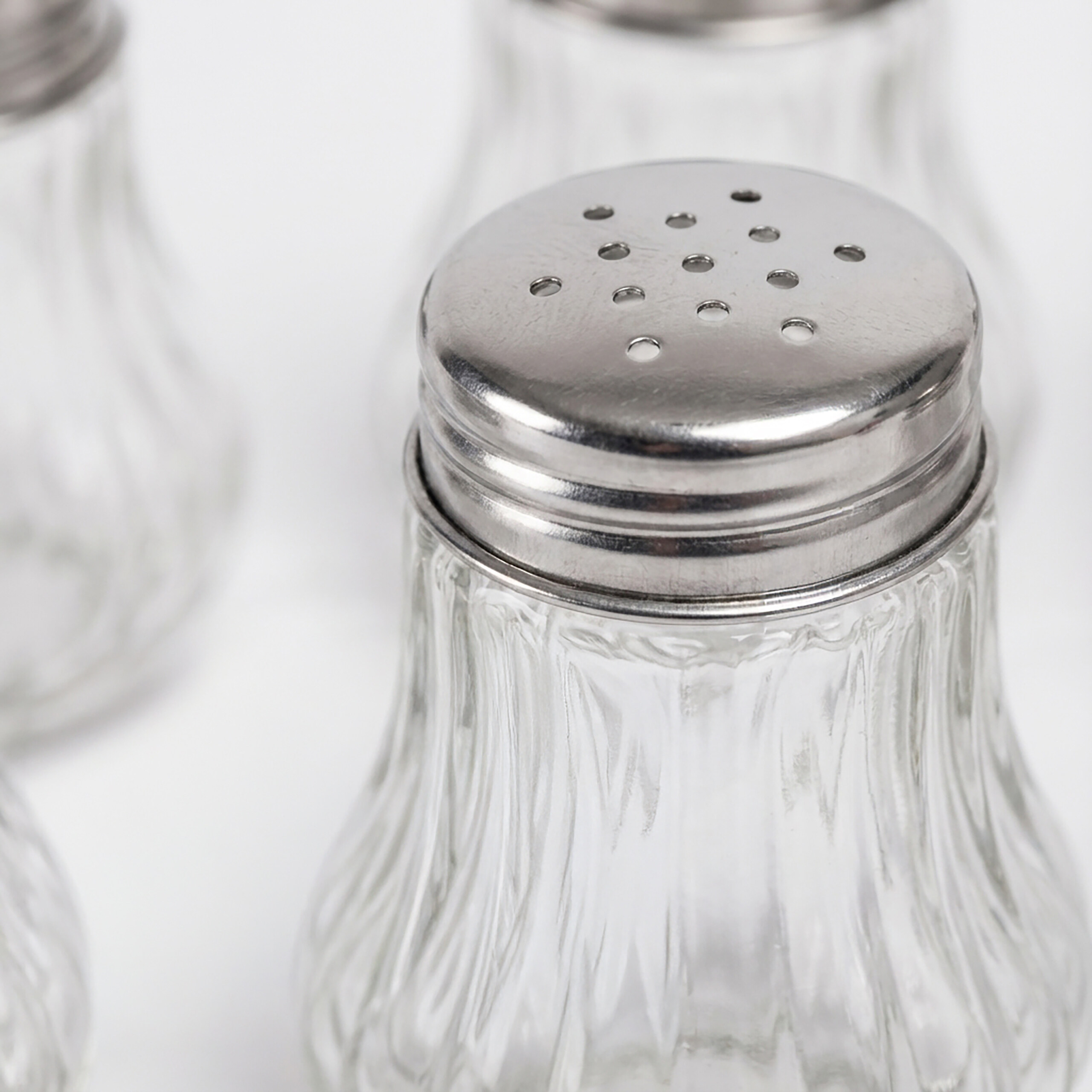 Nahaufnahme eines Salzstreuers aus Glas mit glänzendem Metalldeckel, andere Streuer im Hintergrund unscharf.