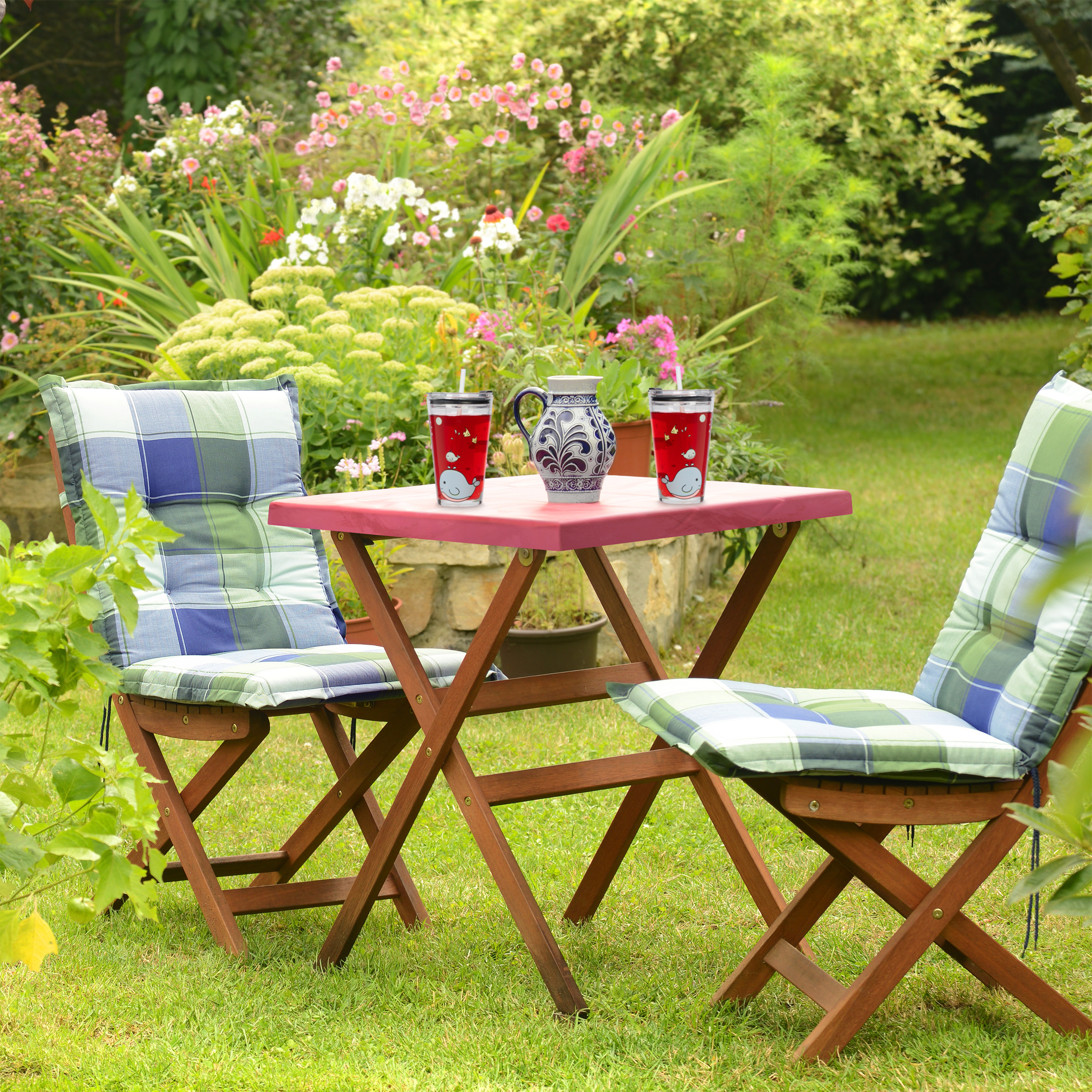Ein Holztisch und zwei karierte Stühle stehen auf einem grünen Rasen in einem Garten.